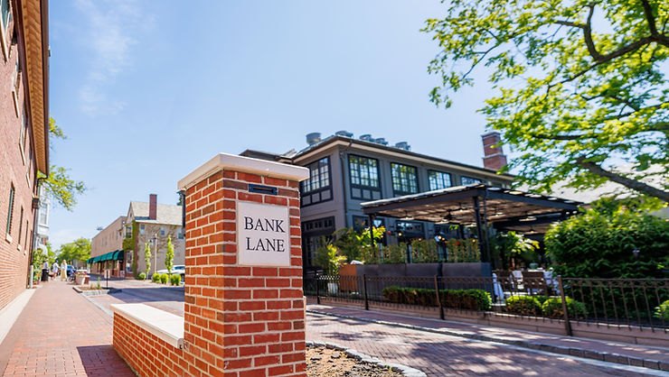Brick pillar with a Bank Lane sign in front of commercial buildings and trees in a downtown setting.