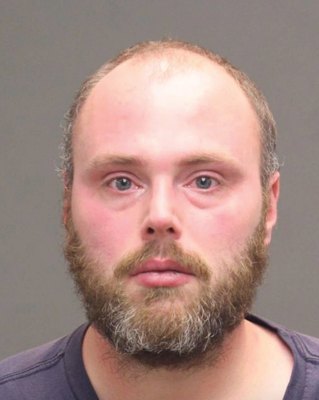 Booking photo of a bearded man in a dark t-shirt against a neutral background.