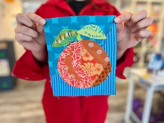 A person in a red hoodie holds up a small fabric collage depicting a fruit with leaves on a blue patterned background.