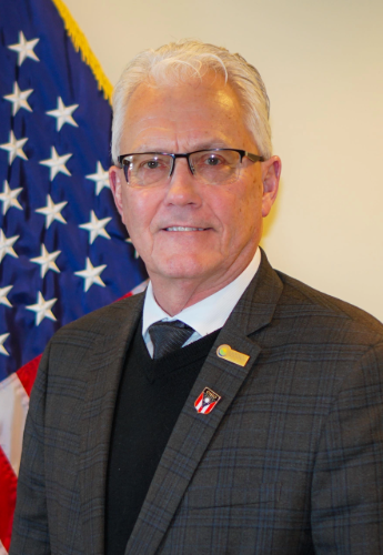 Man with white hair and glasses in a dark blazer stands before an American flag.