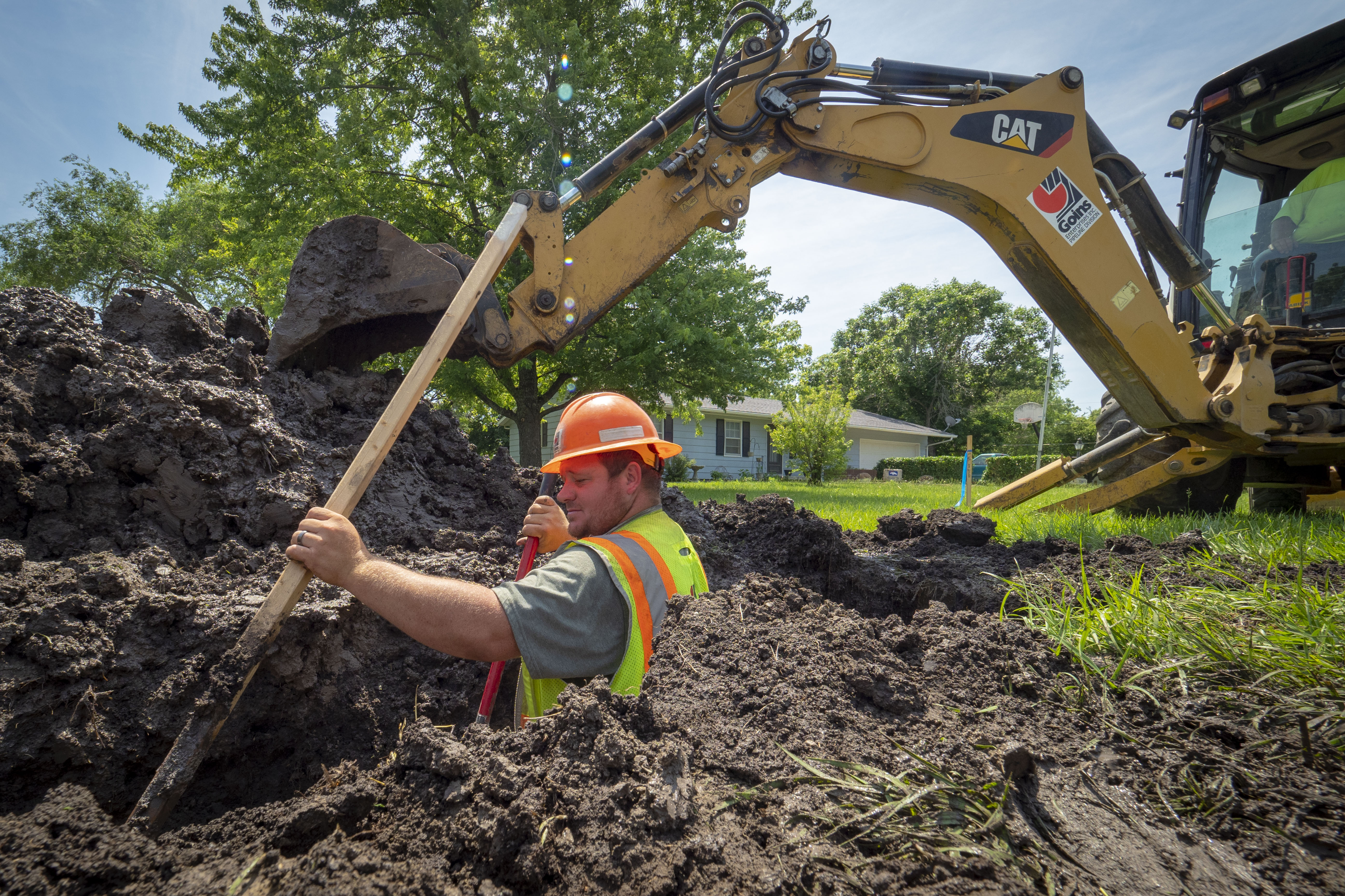 Worker in orange hard hat and safety vest stands in a deep excavation trench holding a shovel.