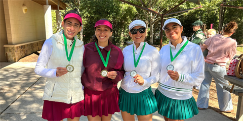 Four women in tennis attire smile and hold medals outdoors.