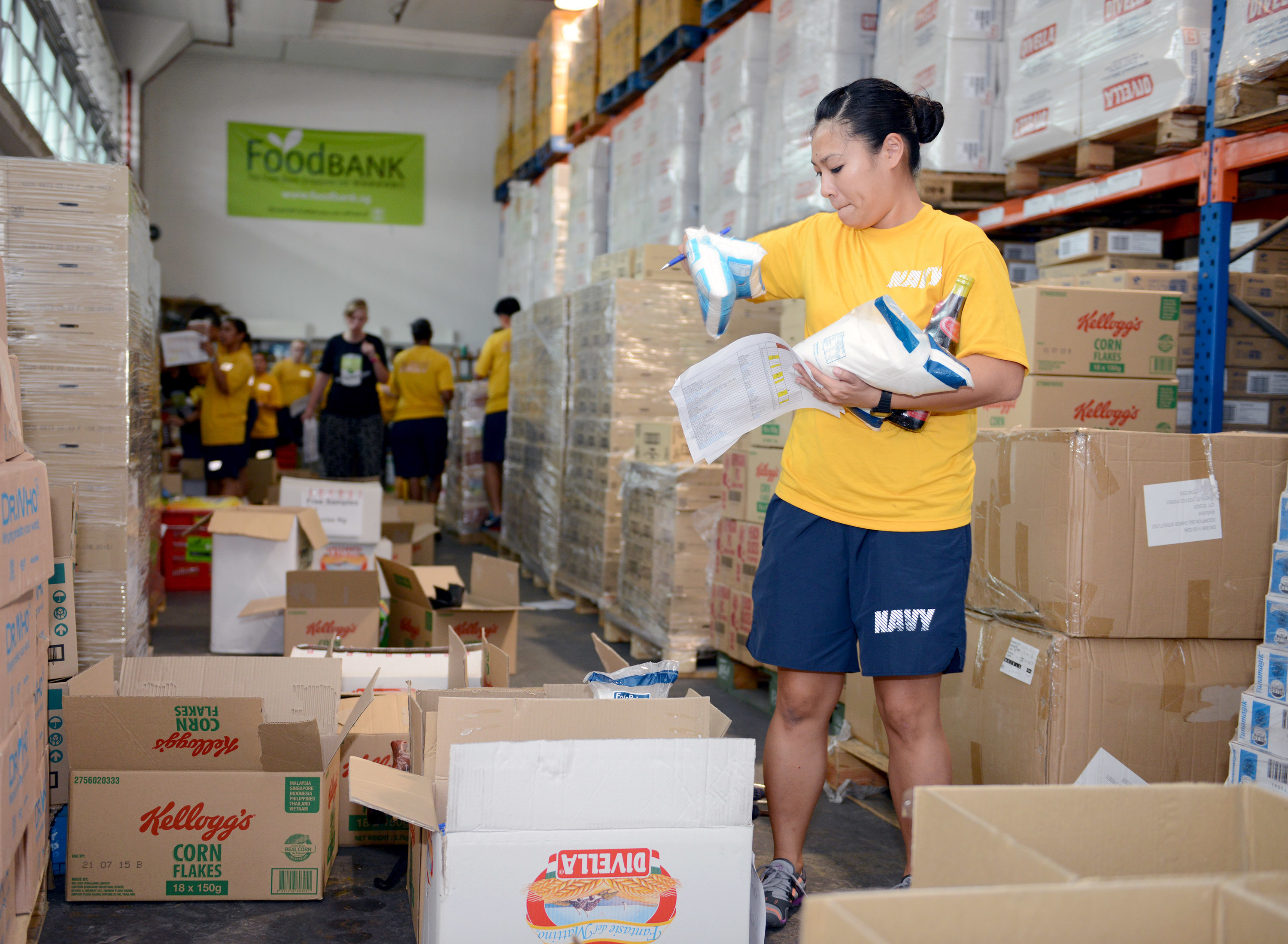 A volunteer in a yellow shirt sorts food items in a warehouse stocked with boxes of dry goods.