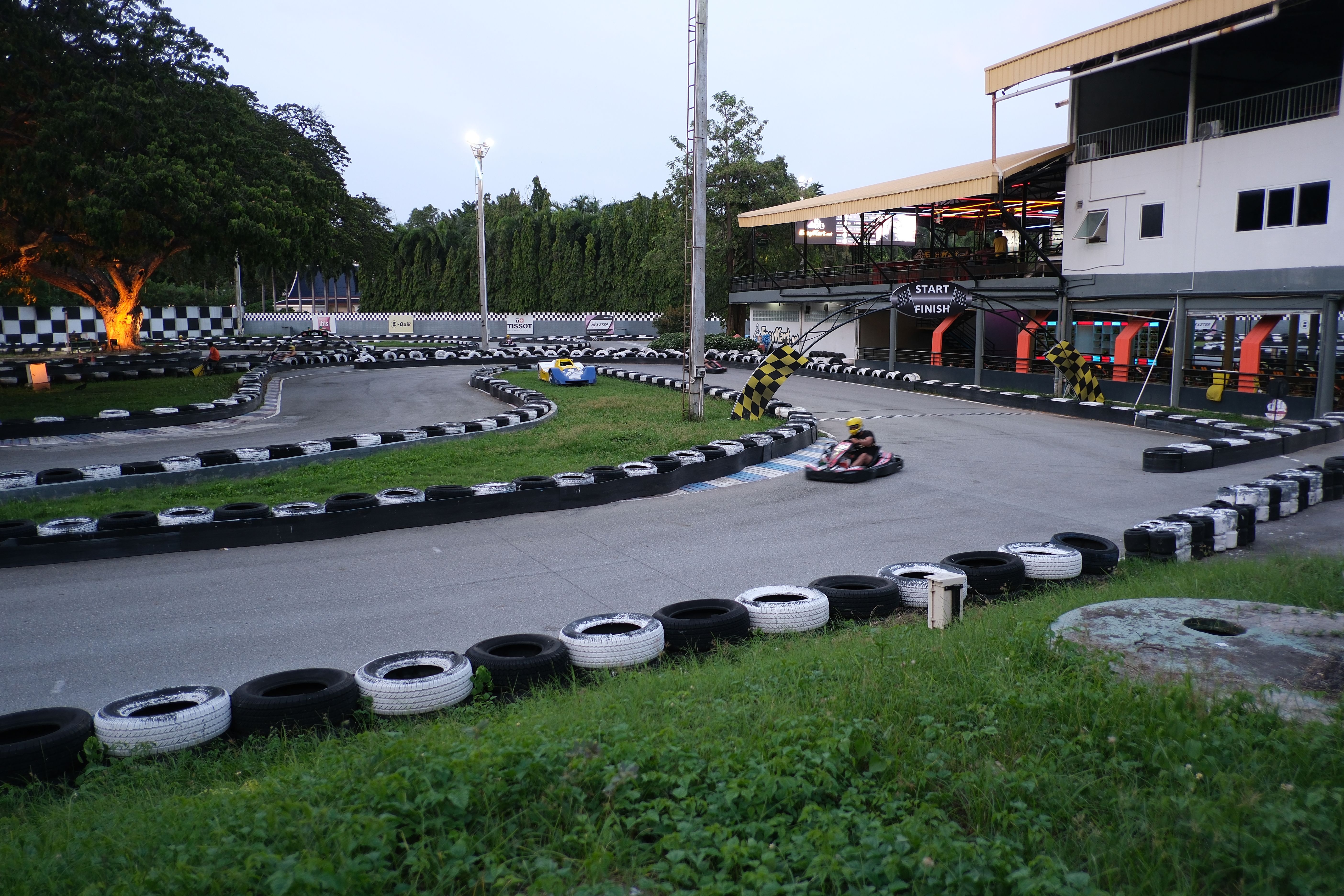 A go-kart races around an outdoor track lined with stacked black and white tires at dusk.
