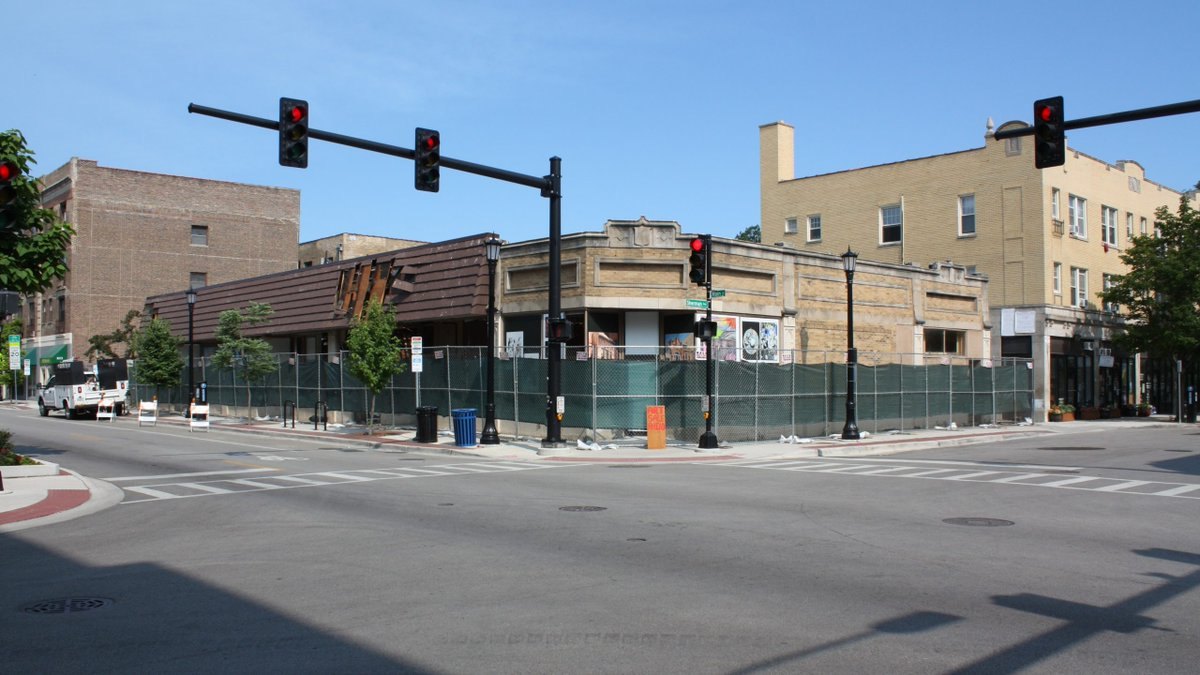 Vacant corner storefront wrapped in green construction fencing at a city intersection.