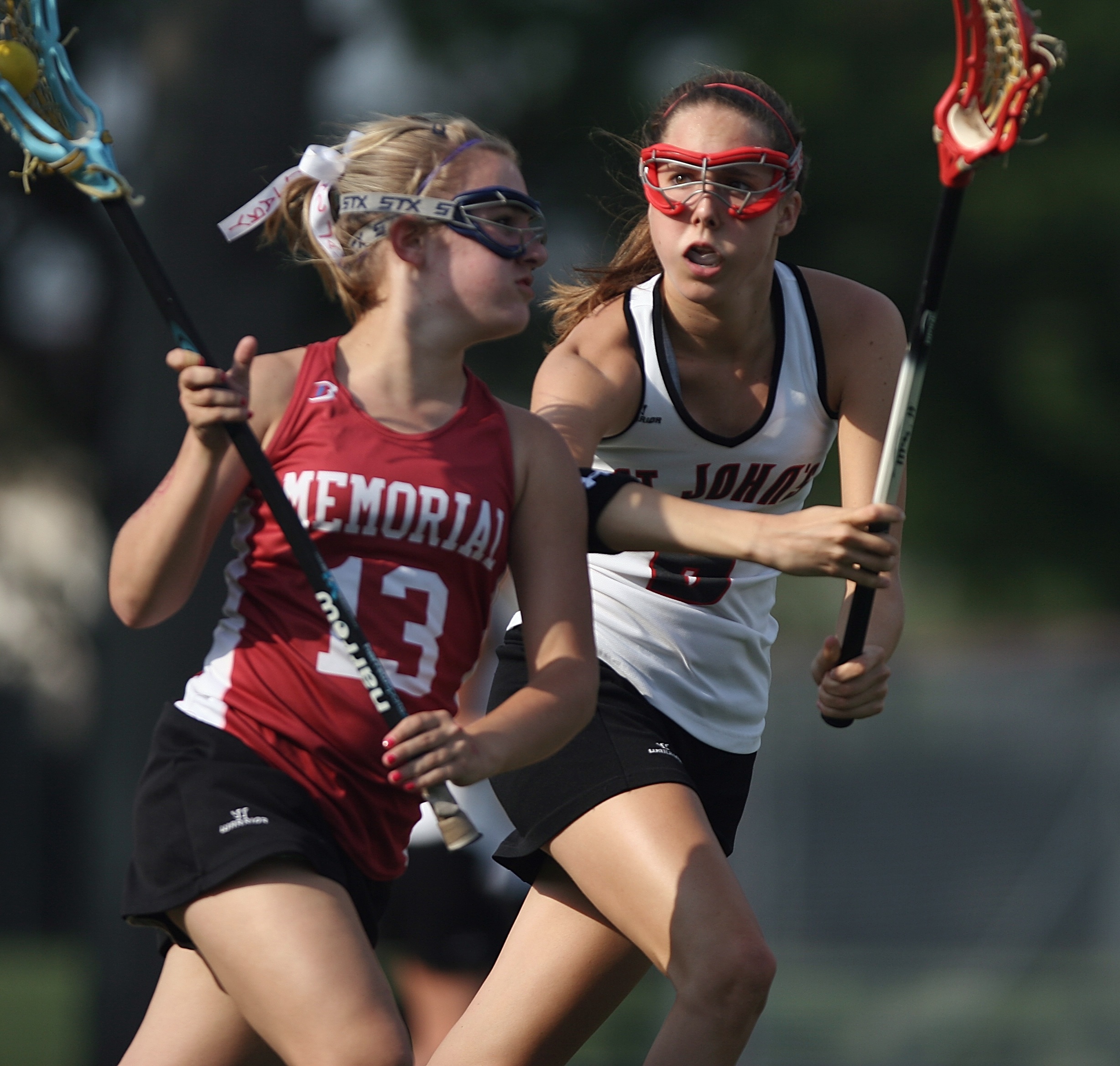 Two female lacrosse players run side by side on a field, each holding a lacrosse stick.
