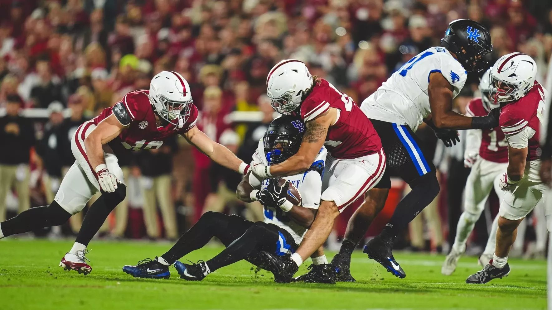 Oklahoma football players in crimson uniforms tackle a Kentucky player in blue on a lit stadium field.