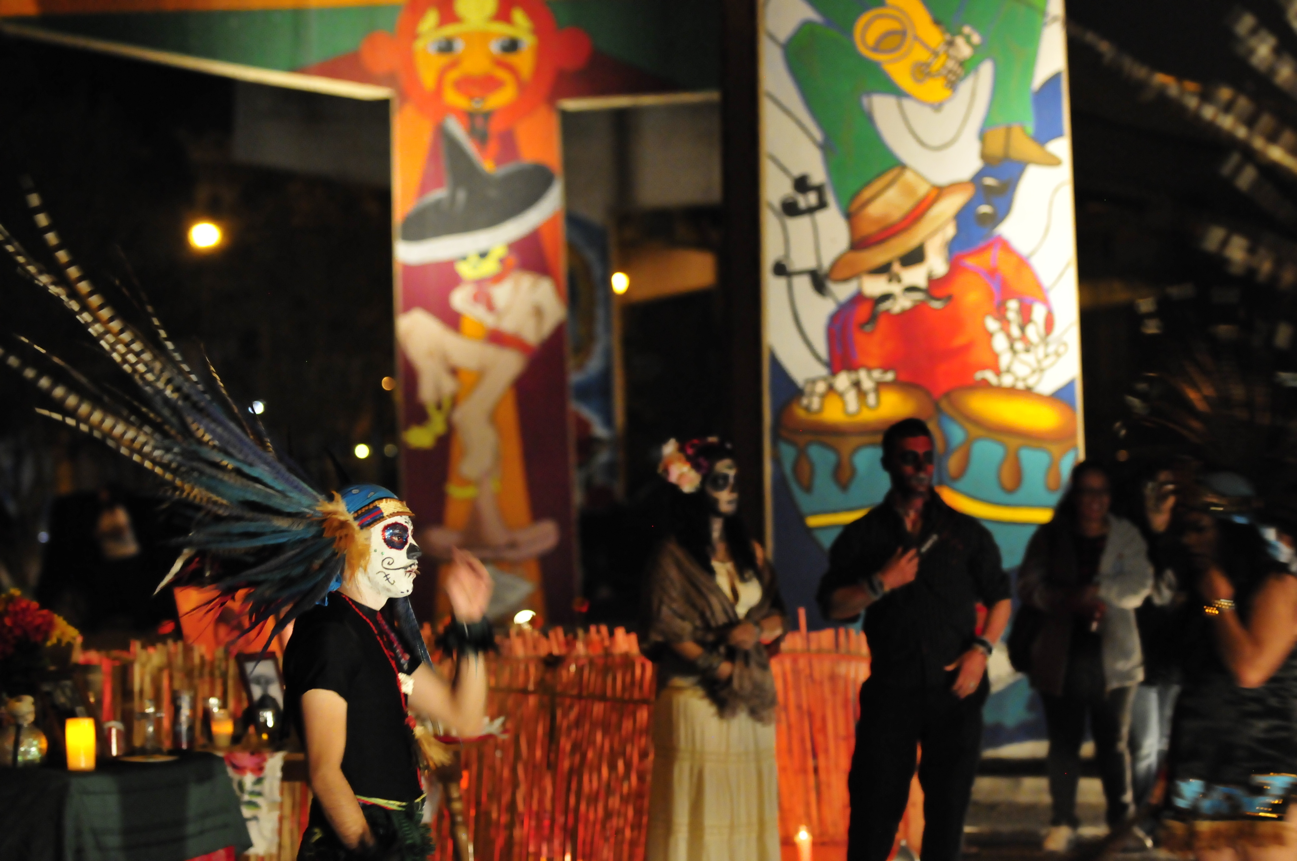 Dancer in Aztec headdress with blue-black feathers and sugar skull face paint at night.