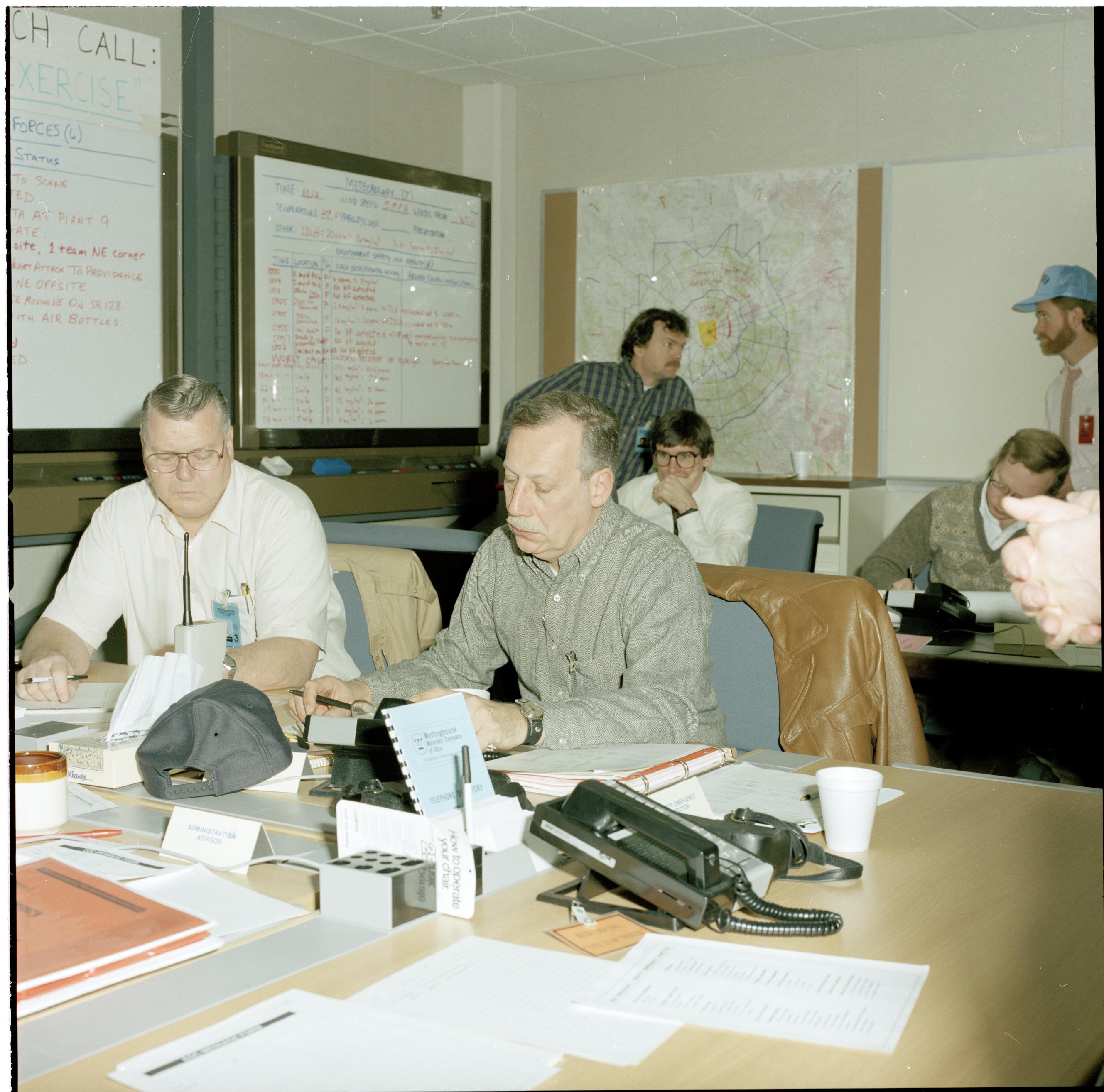 People seated at tables in an emergency operations center with whiteboards and maps visible.