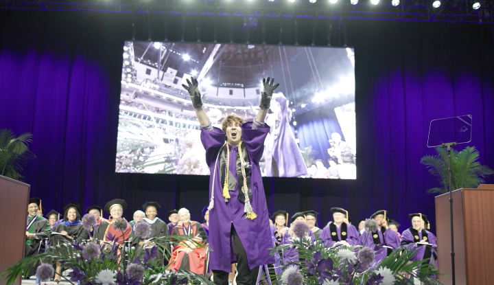 A graduate in purple regalia raises arms in celebration on a commencement stage with a large video screen behind them.
