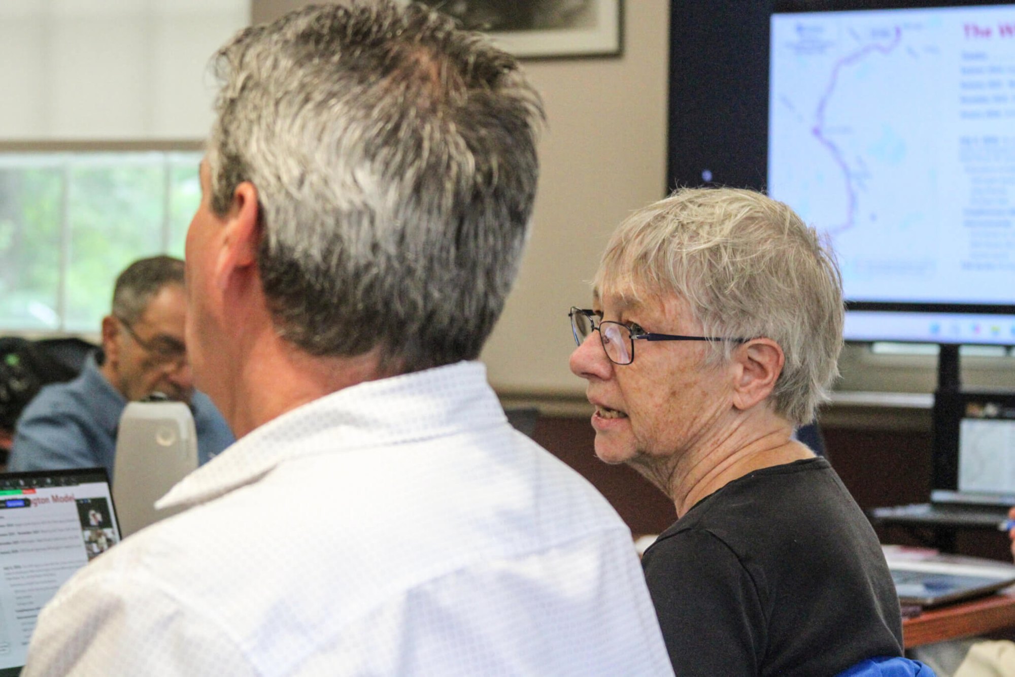 A woman with short white hair and glasses speaks to a man in a meeting room with computer monitors in the background.