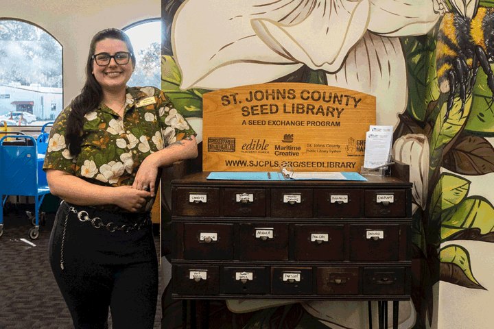 A woman with glasses and a floral shirt stands beside a wooden seed library cabinet in front of a painted floral mural.