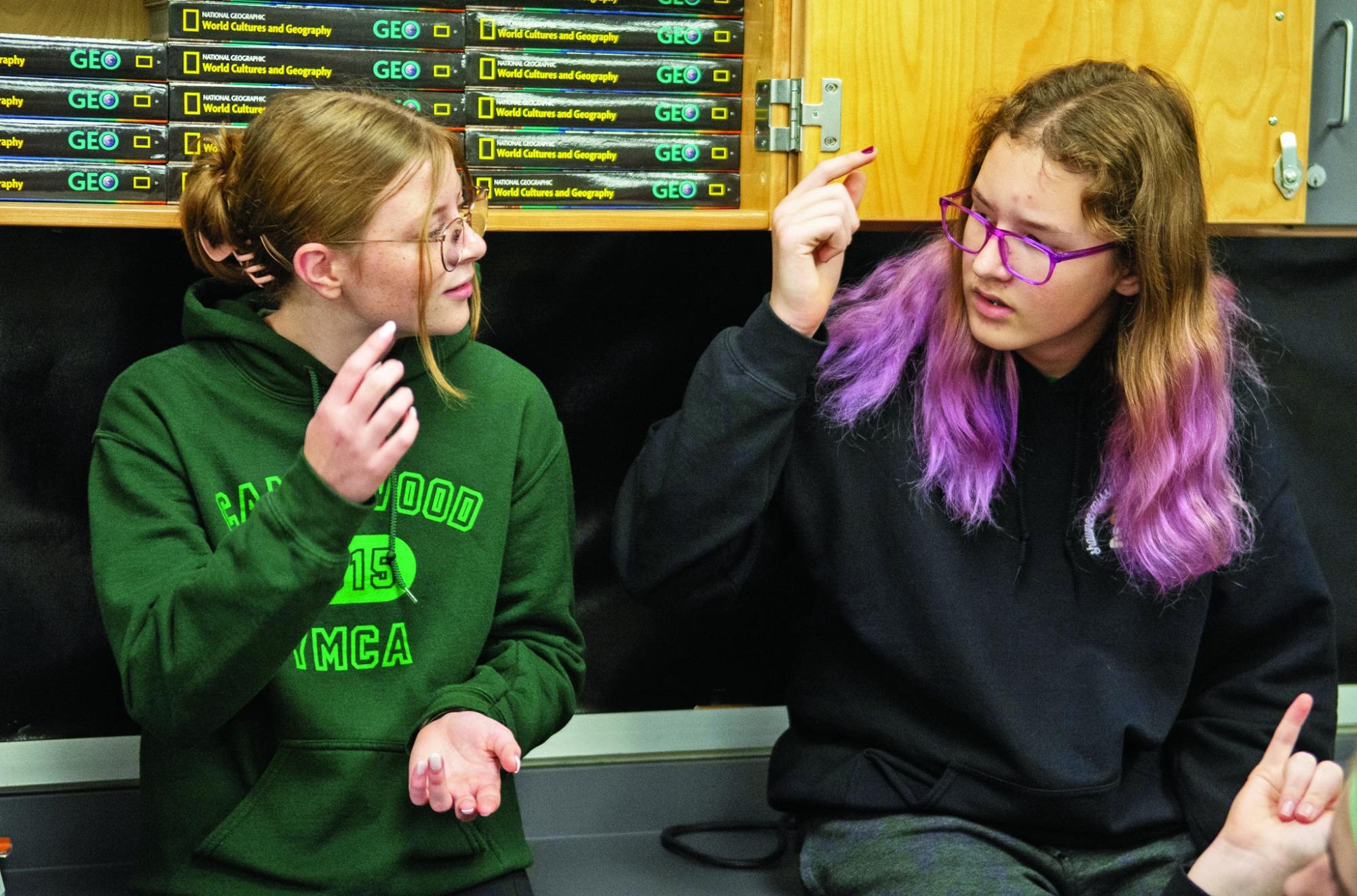 Two students seated in a classroom communicate using hand gestures.