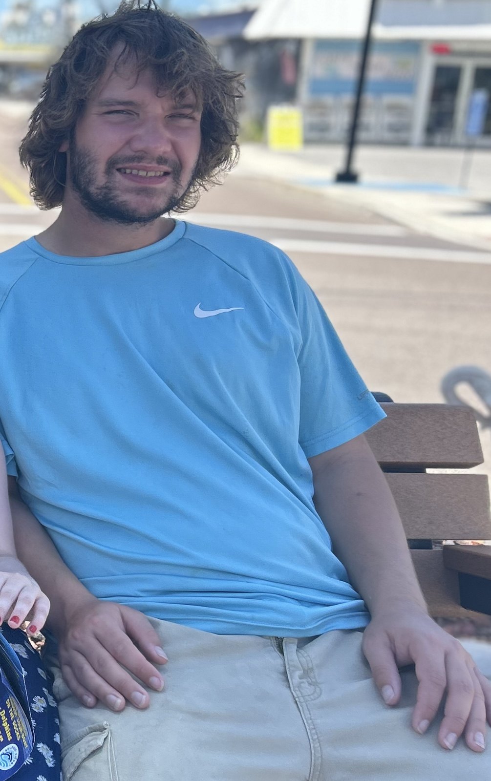Young man with curly brown hair seated on a wooden bench outdoors.
