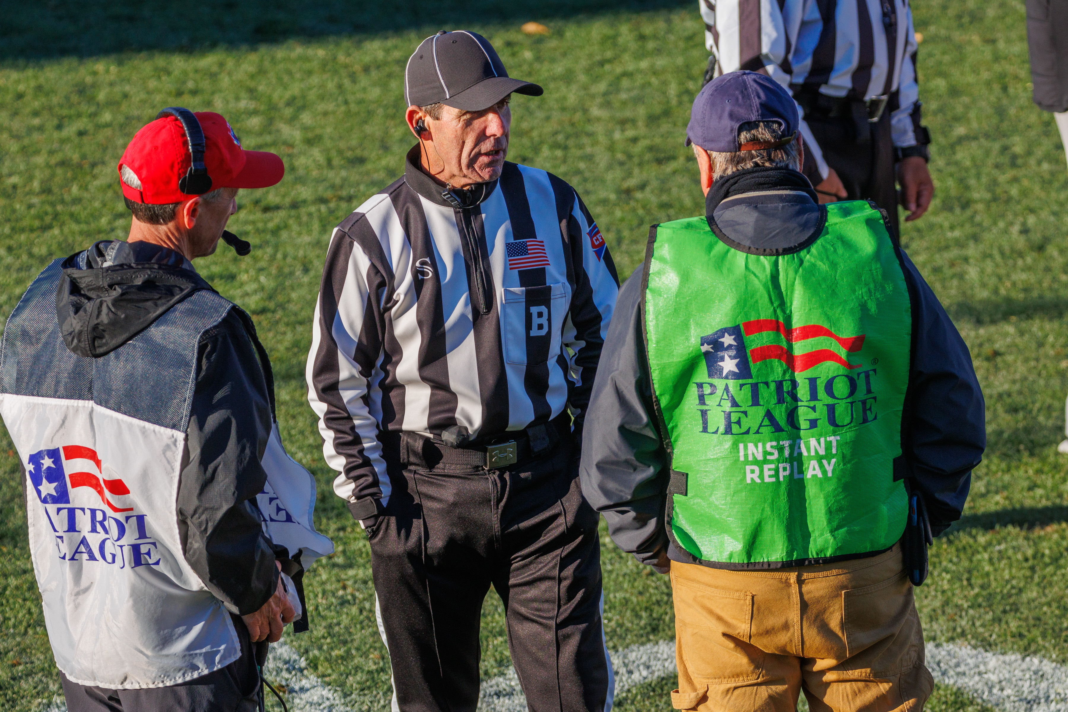 A referee in stripes talks with two officials wearing Patriot League vests on a football field.