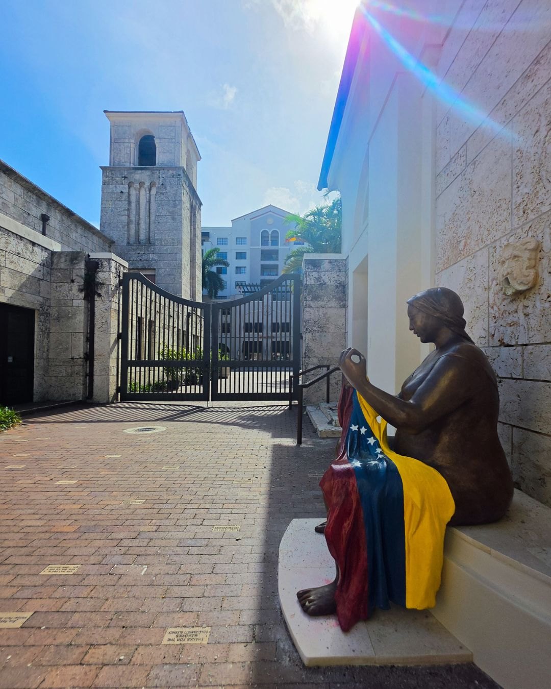 Bronze-colored sculpture of a seated woman stitching a Venezuelan flag, displayed in a courtyard with a stone bell tower in the background.