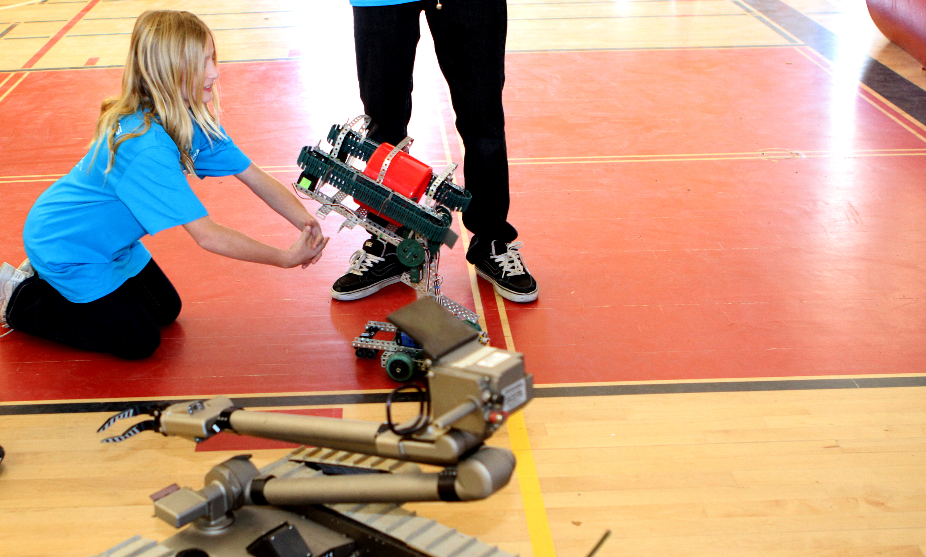 A student kneels on a gym floor working with a small robot while another student stands nearby.