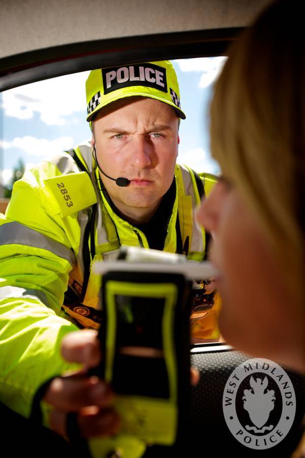 Police officer in yellow jacket holds a testing device toward a car window.