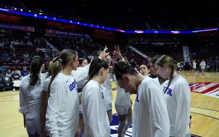 Women's basketball players in white warm-up shirts huddle together on a hardwood court.