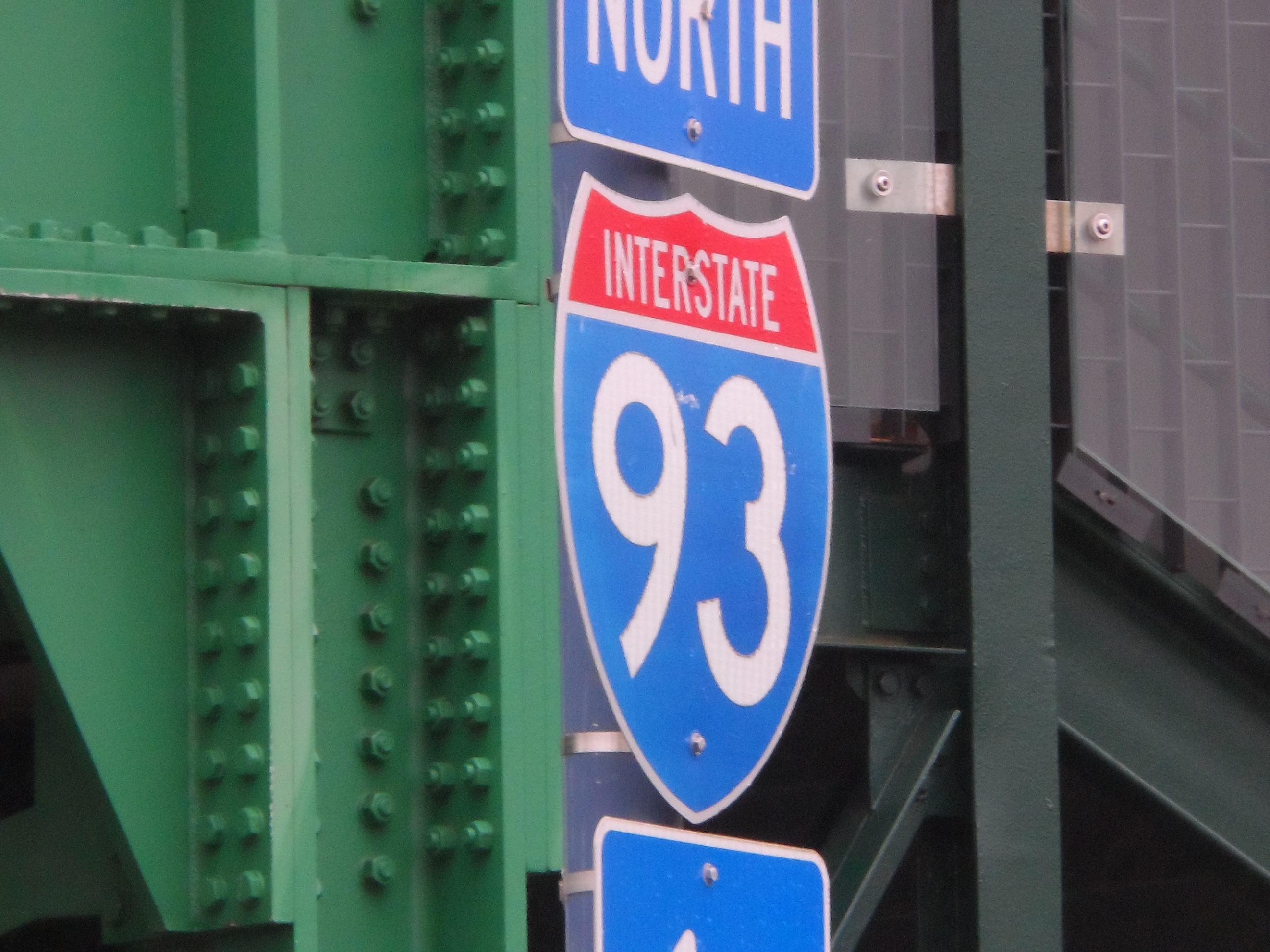 Blue and red Interstate 93 North shield sign mounted on a green steel bridge.