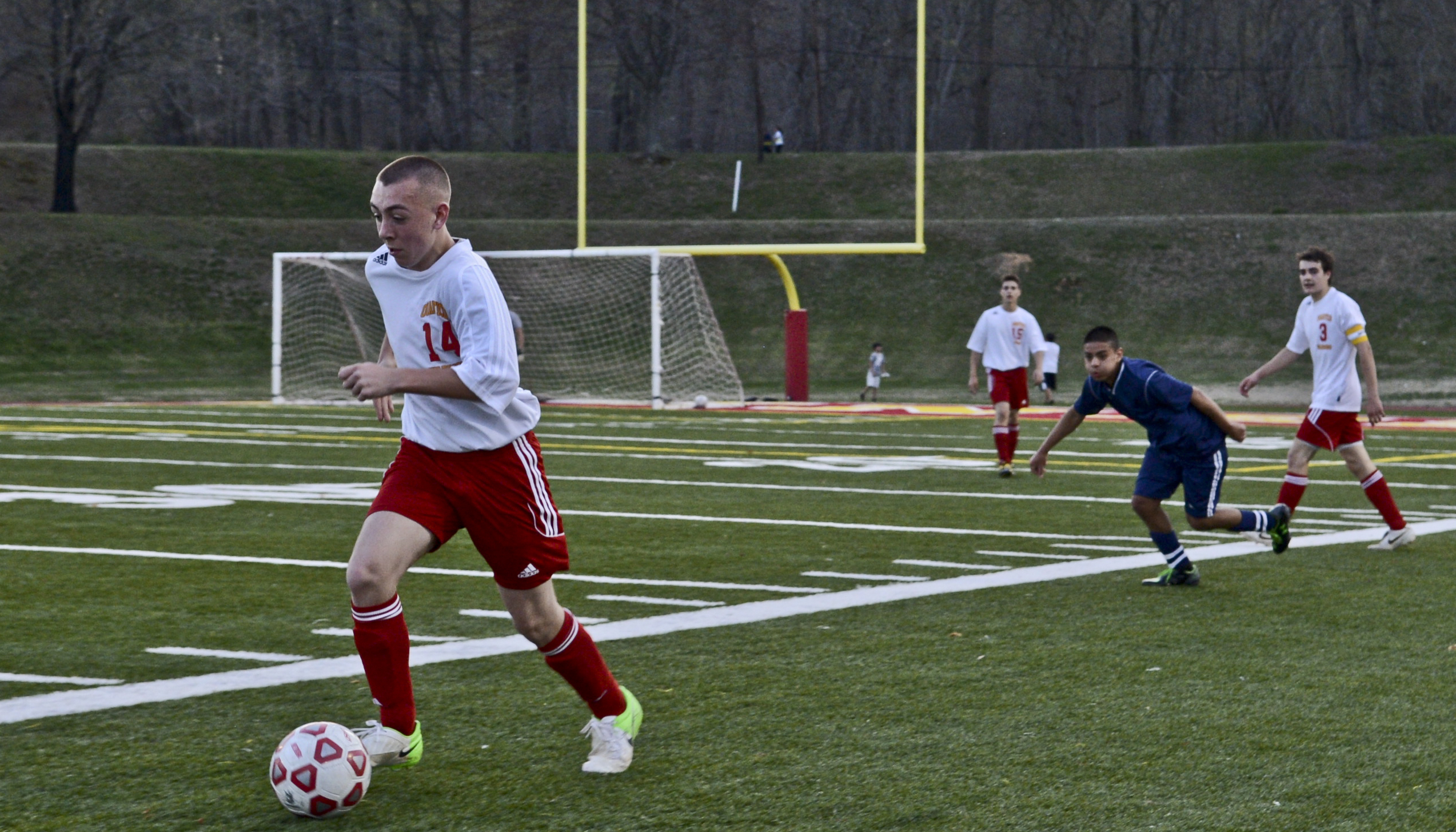 A soccer player in a white jersey dribbles a ball down a green turf field.