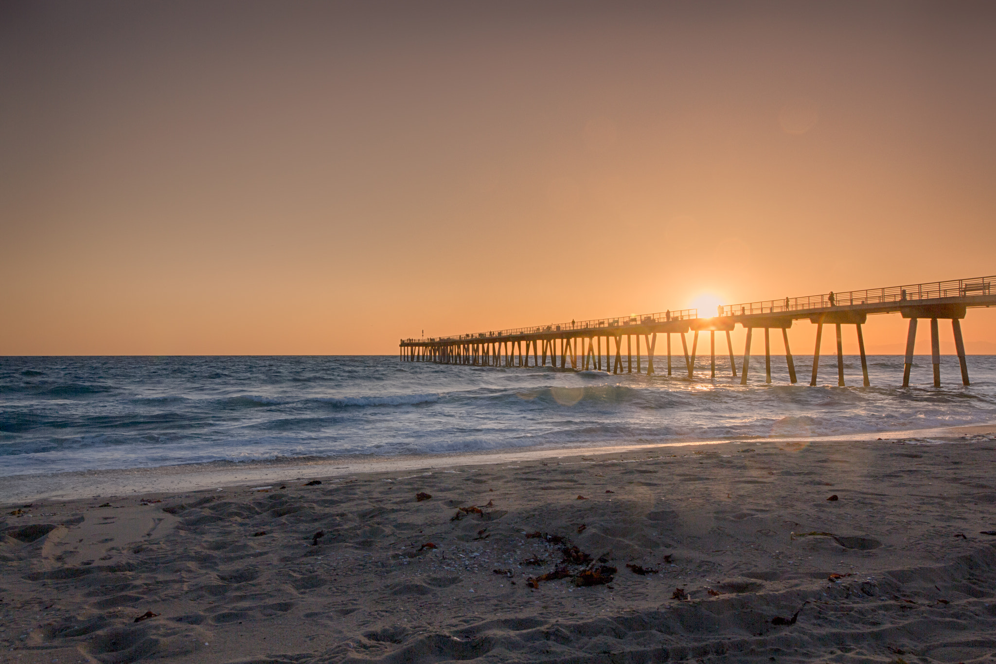 Long wooden pier extending over the ocean at golden sunset with sandy beach in the foreground.