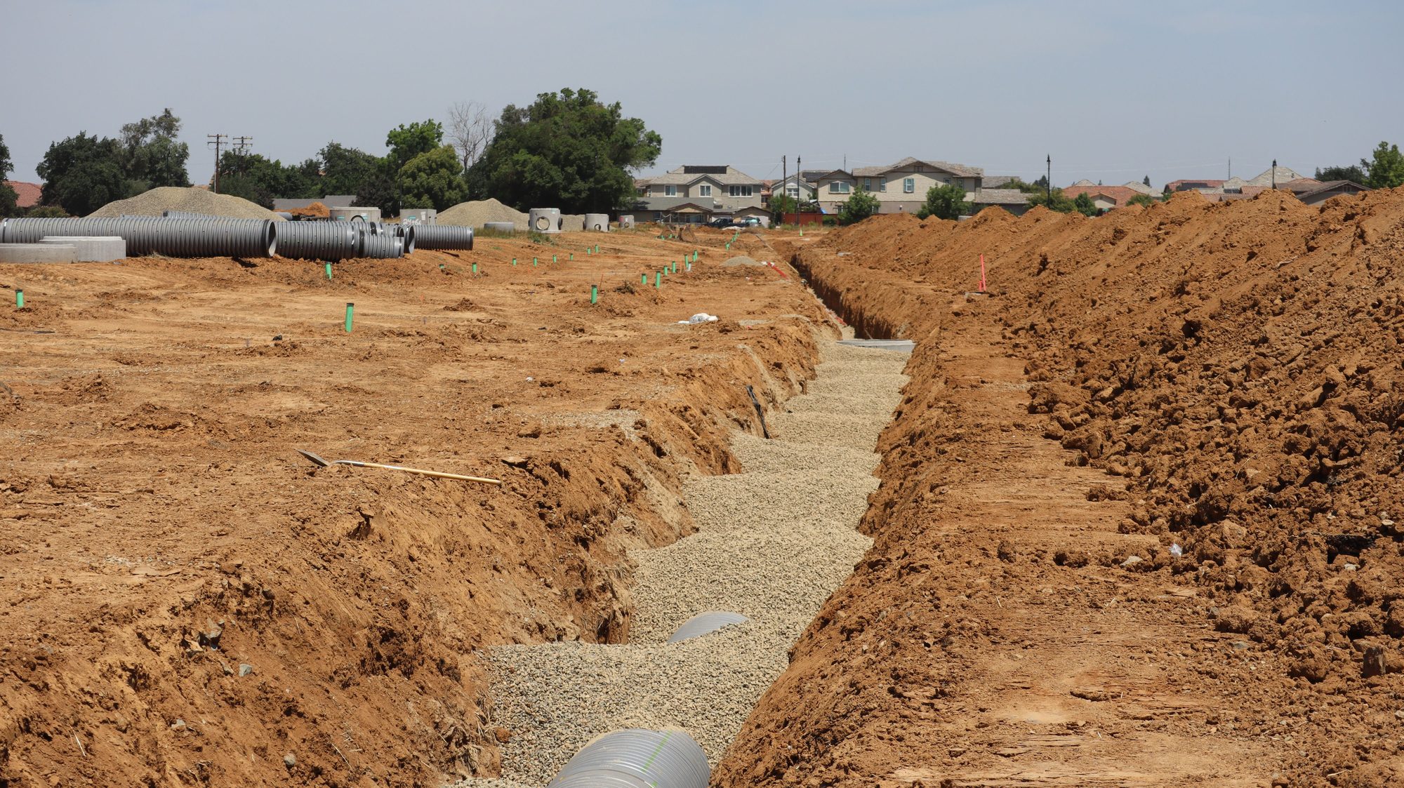 Wide view of a construction site with trenches and large pipes in a suburban setting under a clear sky.