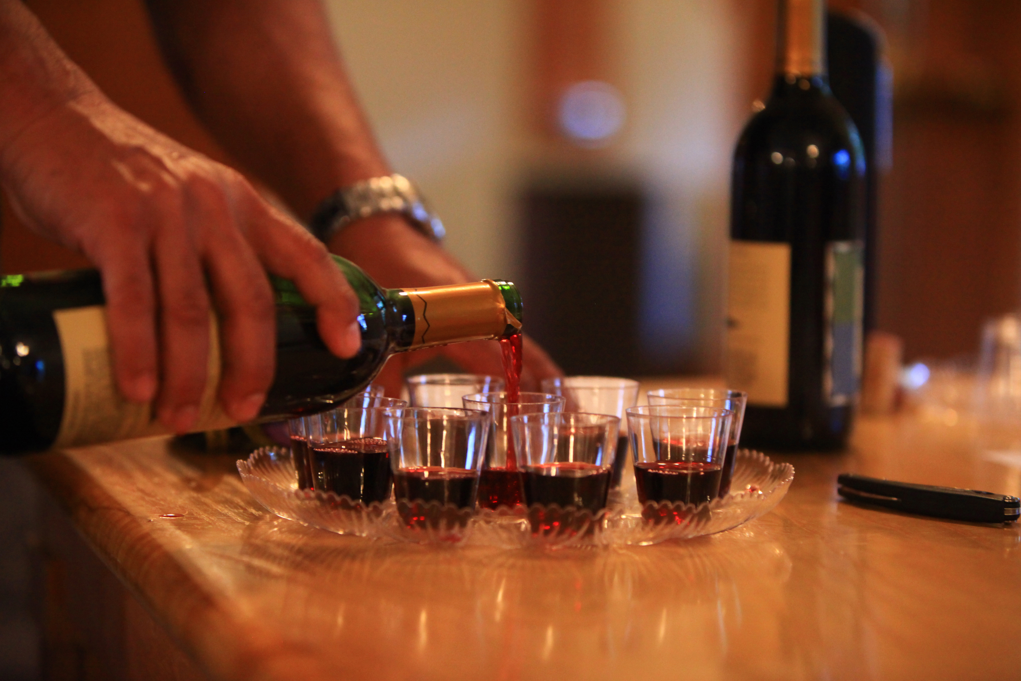 A hand pours red wine from a green bottle into small tasting glasses on a wooden surface.