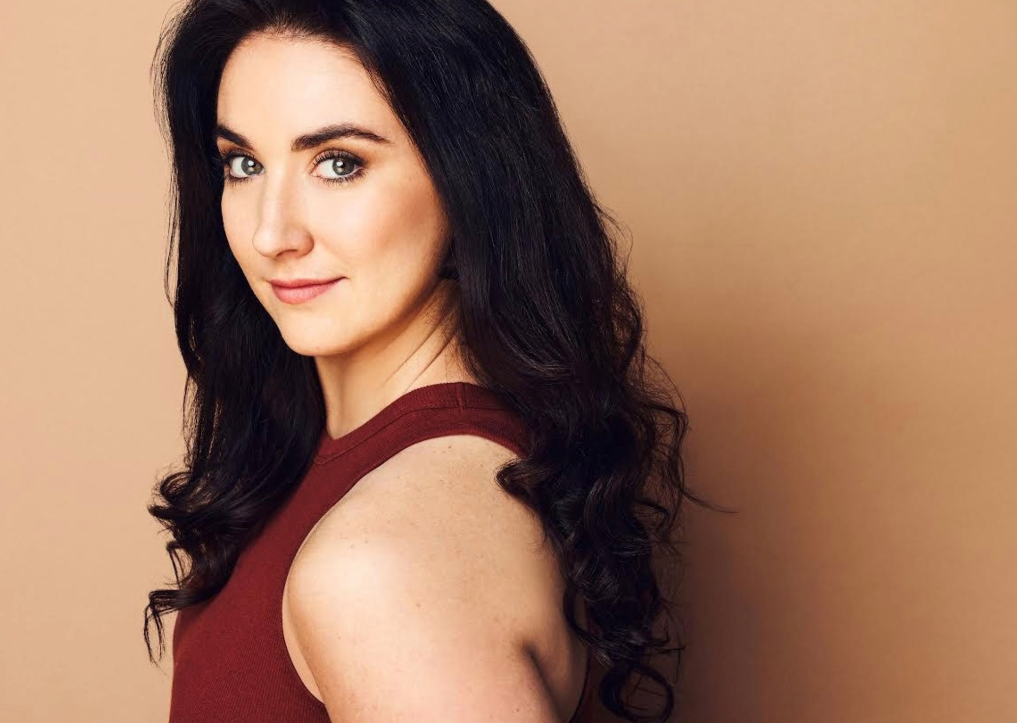 Woman with long dark curly hair in a sleeveless dark red top posing for a professional headshot.