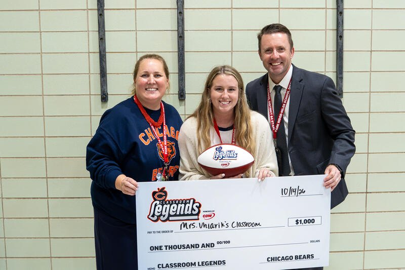 Three adults pose indoors; the center person holds a large ceremonial check and a football.