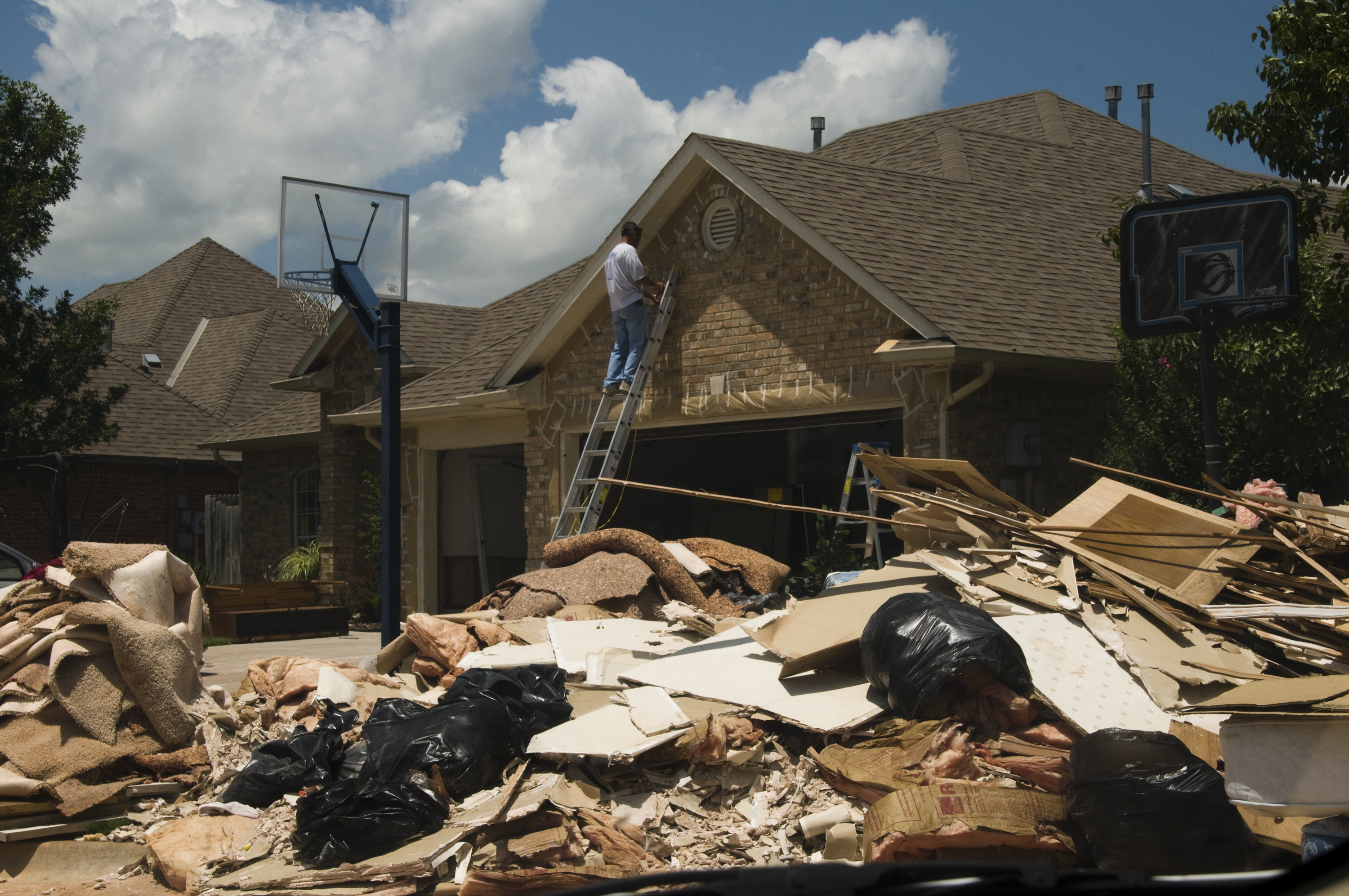 Homeowner on a ladder works on a brick house surrounded by large piles of storm debris.