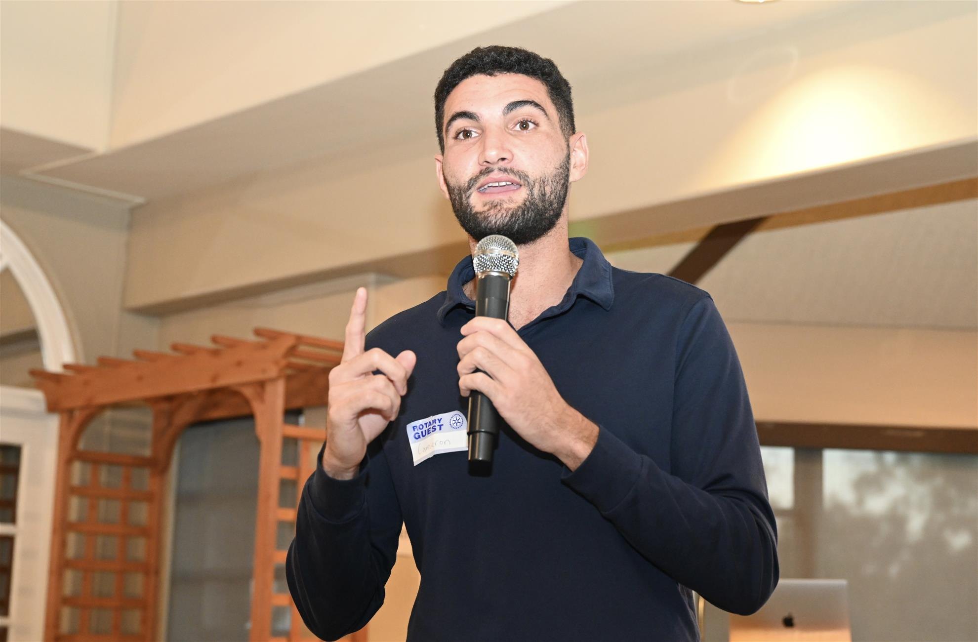 Young bearded man in a navy polo shirt speaks into a microphone at an indoor event.