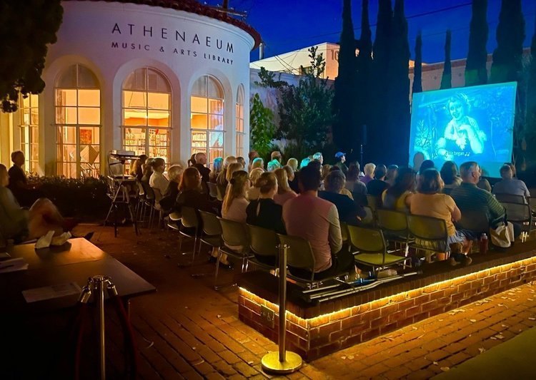 Crowd seated outdoors at night watching a film projected on a large screen, with a lit library building in the background.