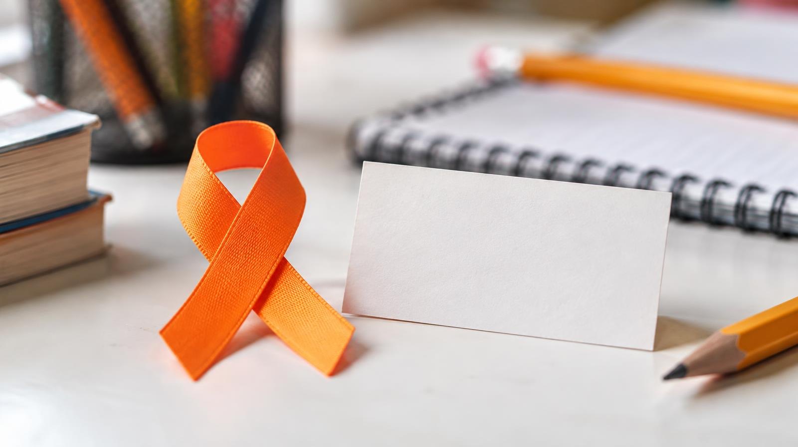 Orange awareness ribbon beside pencils and a notebook on a white surface