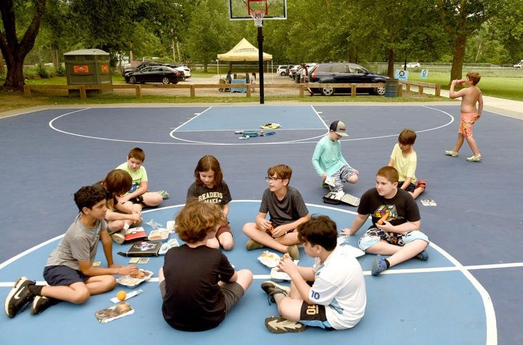 Children seated in a circle on a basketball court eating free lunches outdoors.