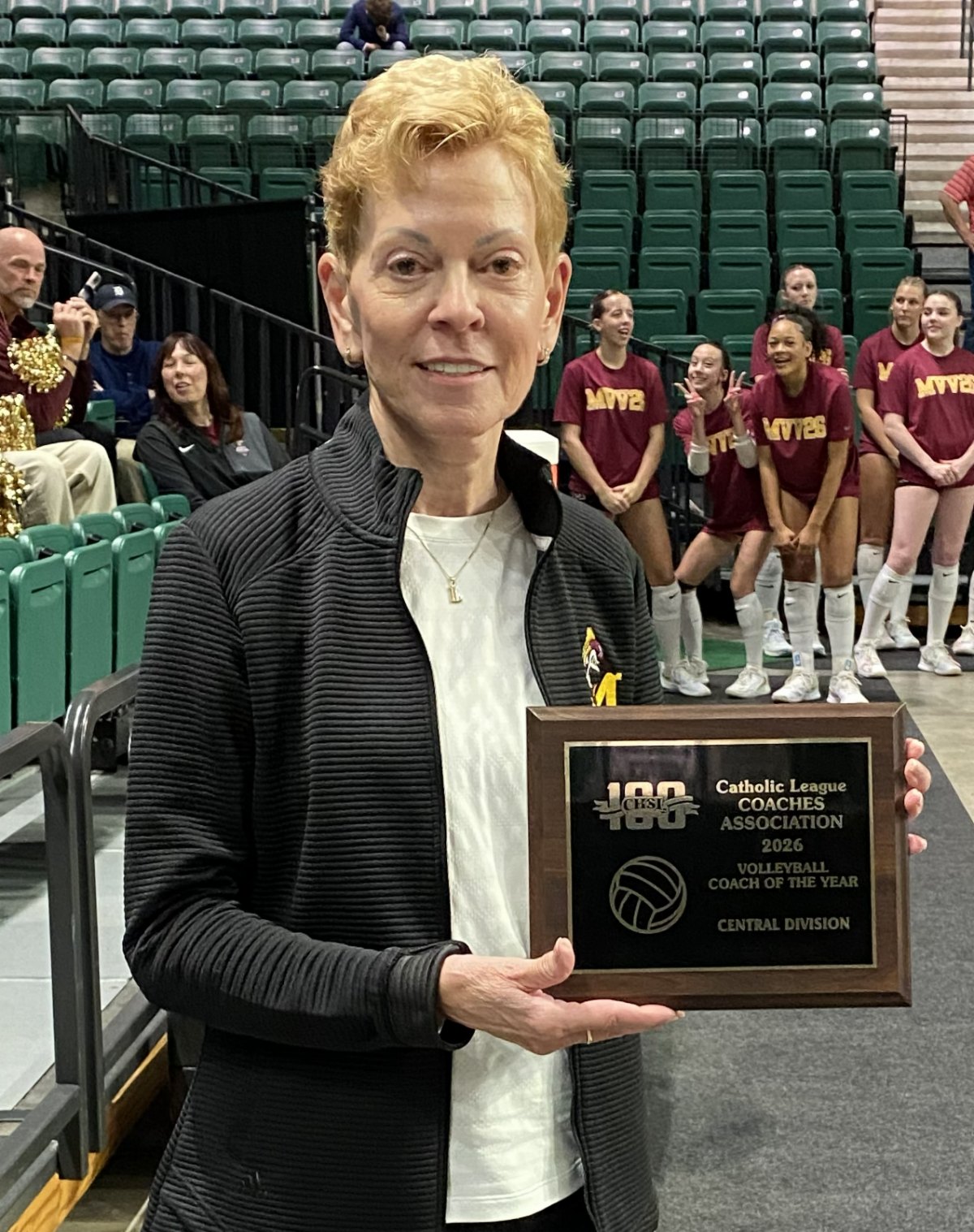 A woman holds a wooden Coach of the Year plaque inside a gymnasium, with volleyball players in maroon uniforms behind her.