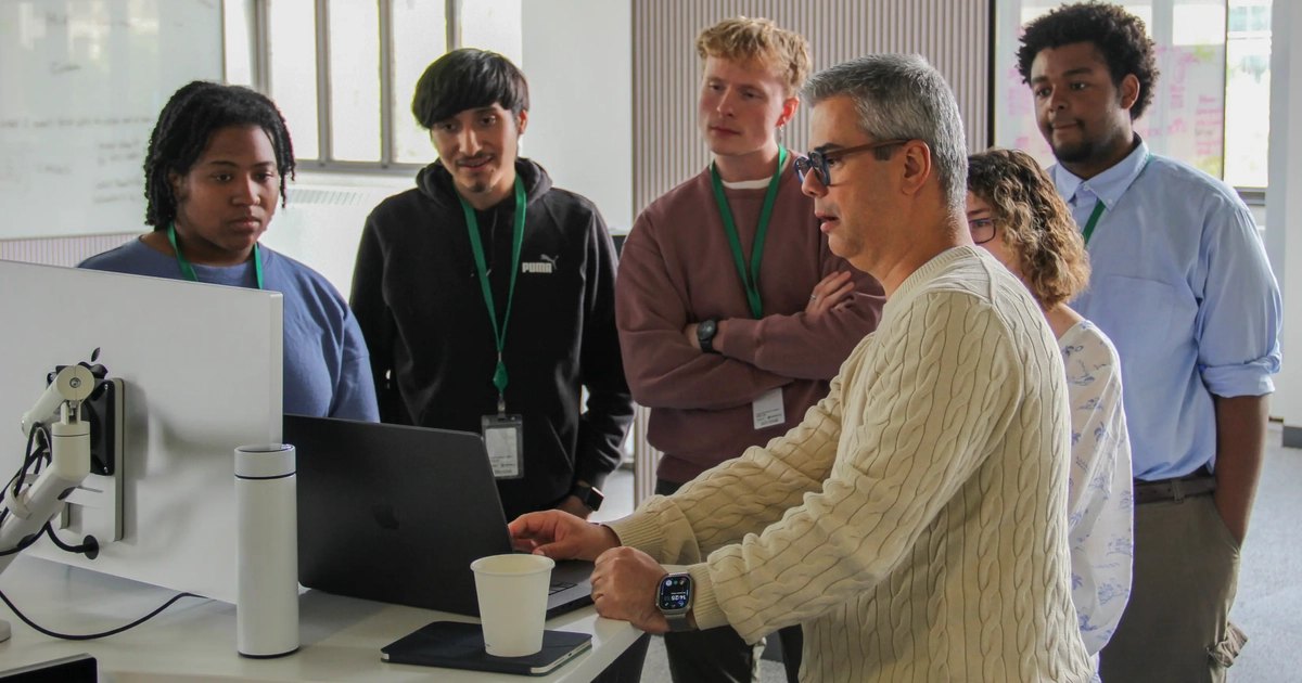Man in cream sweater points at laptop screen while five students with lanyards look on.