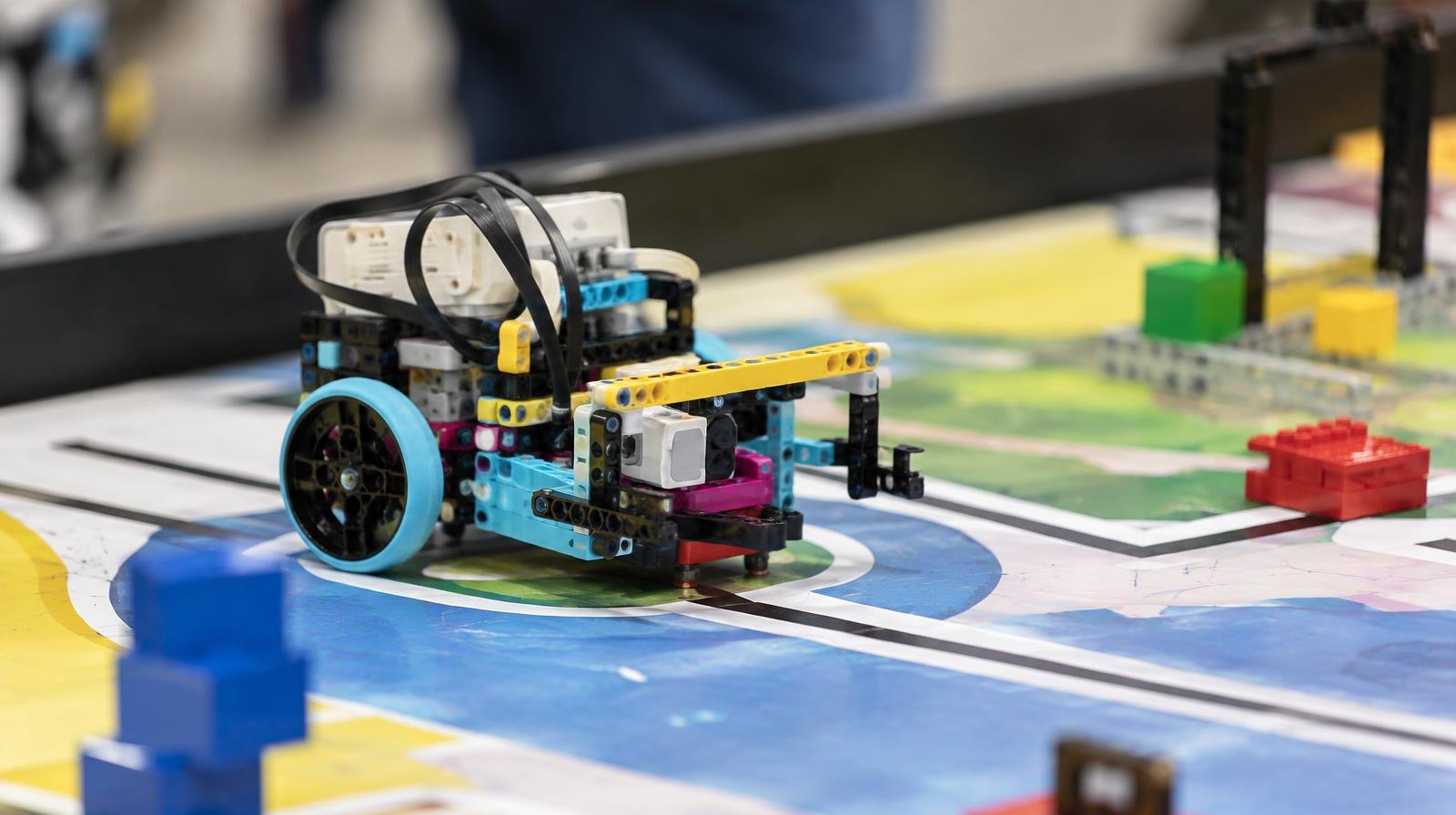 A colorful brick-built robot rests on a printed competition mat under bright lights.