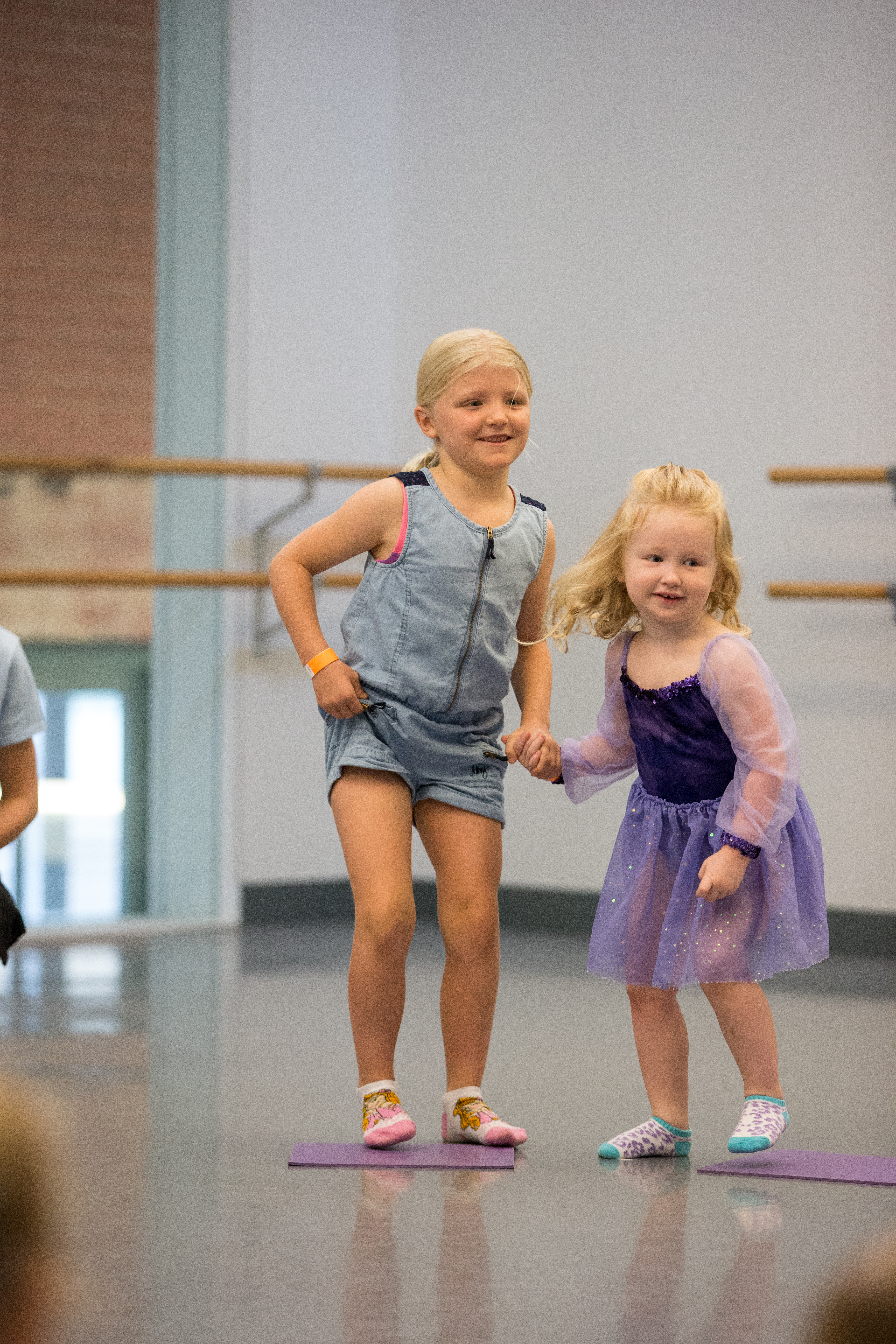 Two young girls in a dance studio smile and hold hands near a ballet barre.