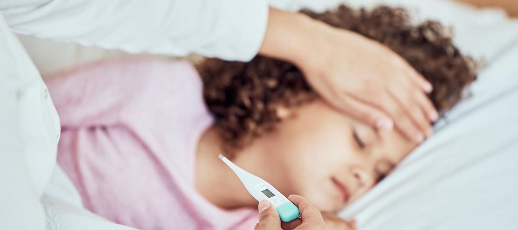 Young child lying in bed with eyes closed while an adult checks forehead and holds a thermometer.