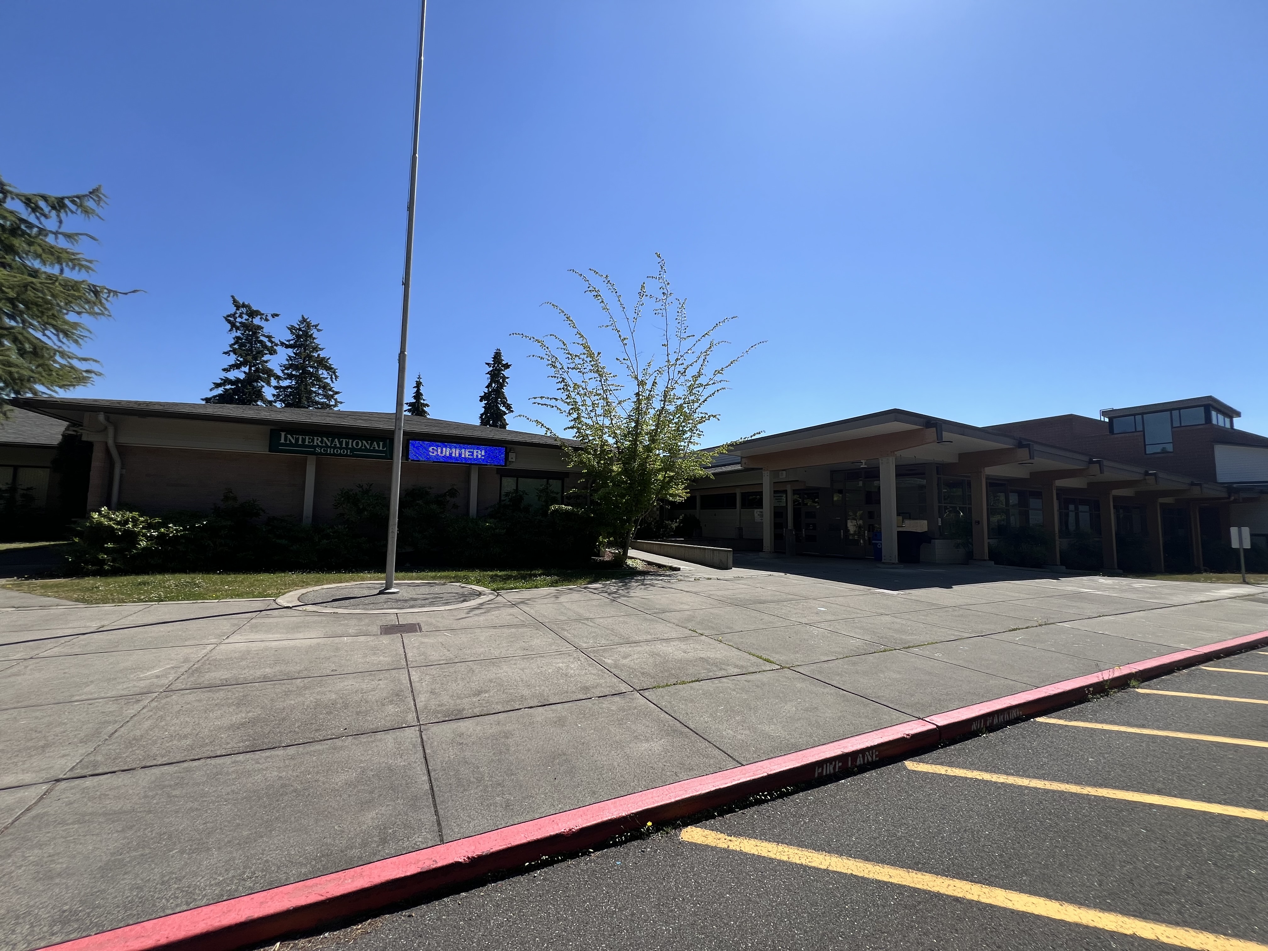 Single-story school building entrance with flagpole and empty parking lot on a sunny day.
