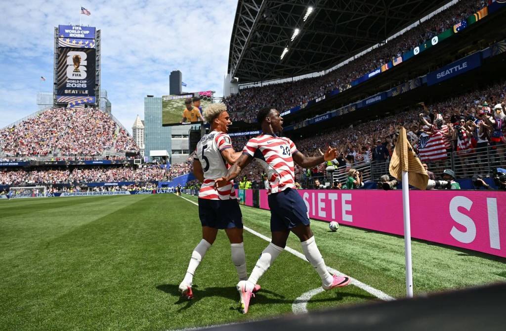 Two USMNT players in red-and-white jerseys celebrating near a corner flag in a packed stadium.