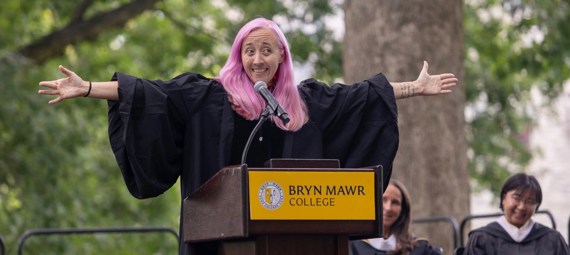 A pink-haired speaker in academic robes stands at a podium with arms outstretched.