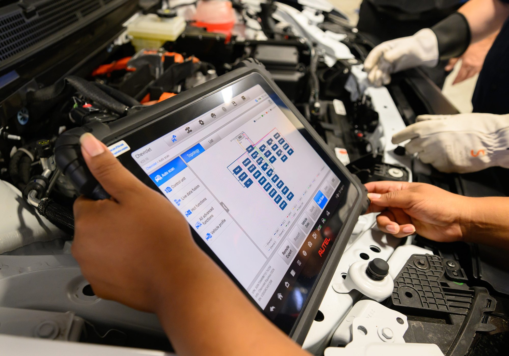 Person holds a diagnostic tablet over an open vehicle engine bay in an automotive shop.
