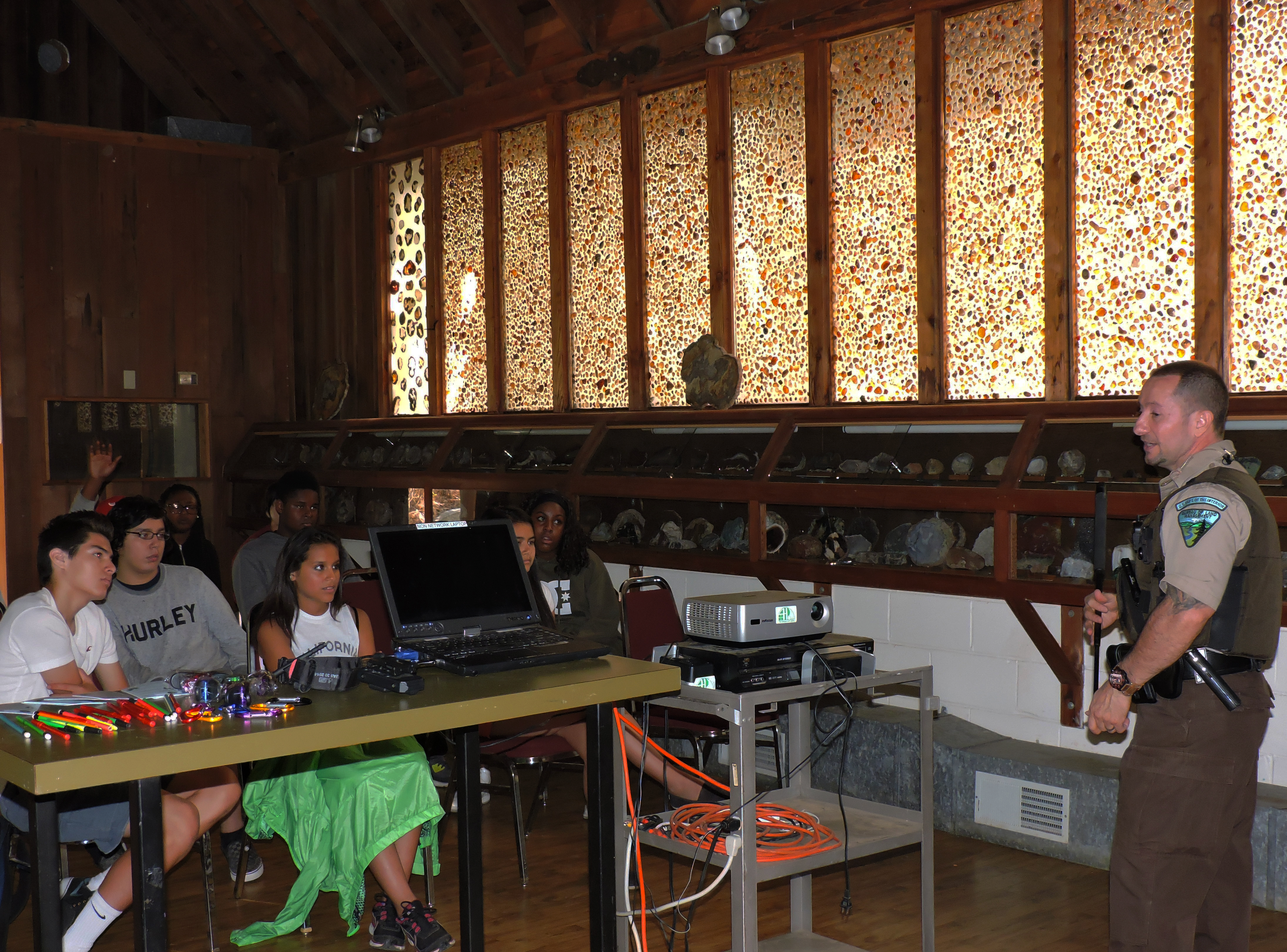 Uniformed officer presenting to students seated at tables in an indoor room.