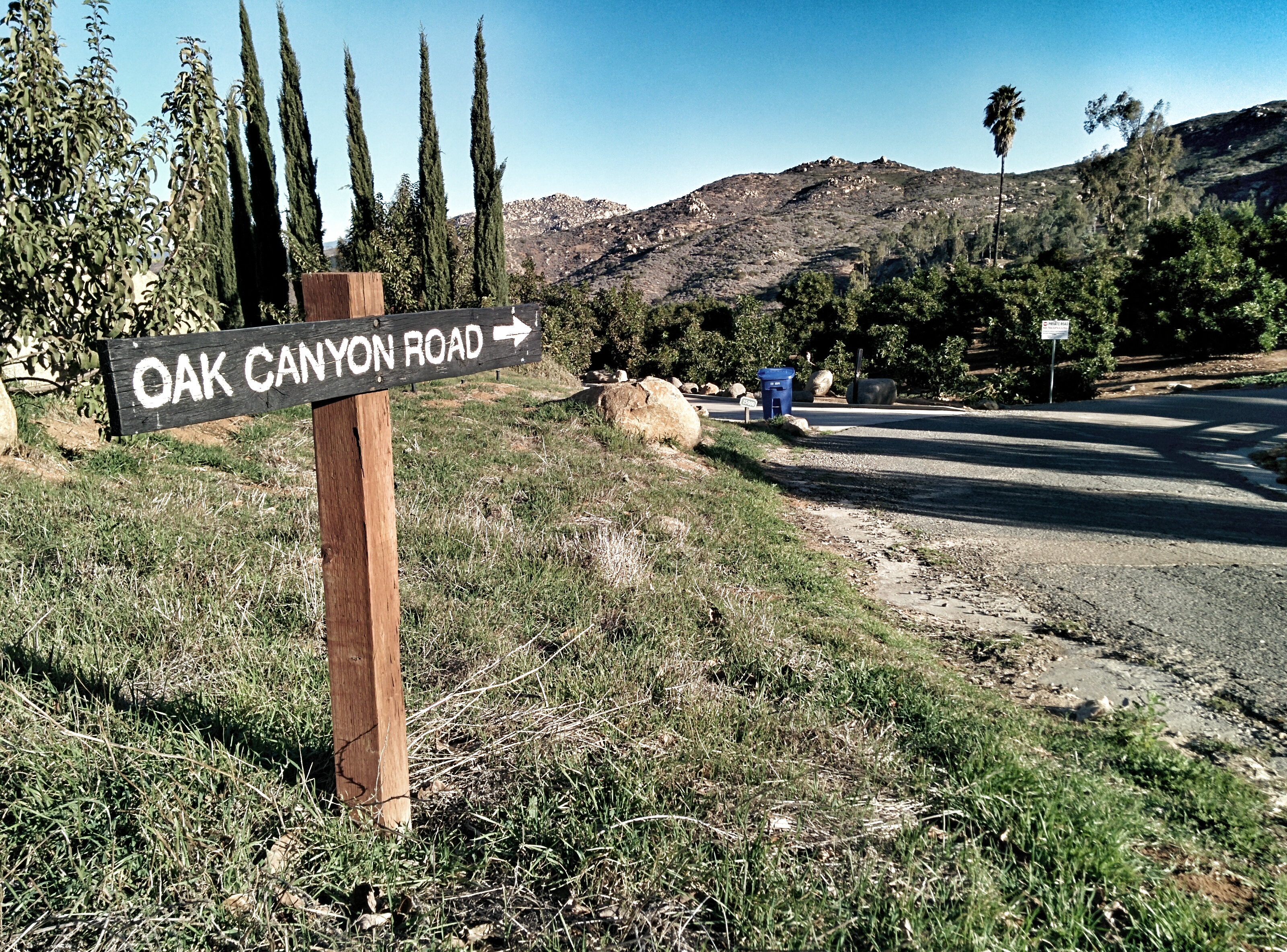 Wooden road sign on a post beside a paved road with hills and trees behind it.