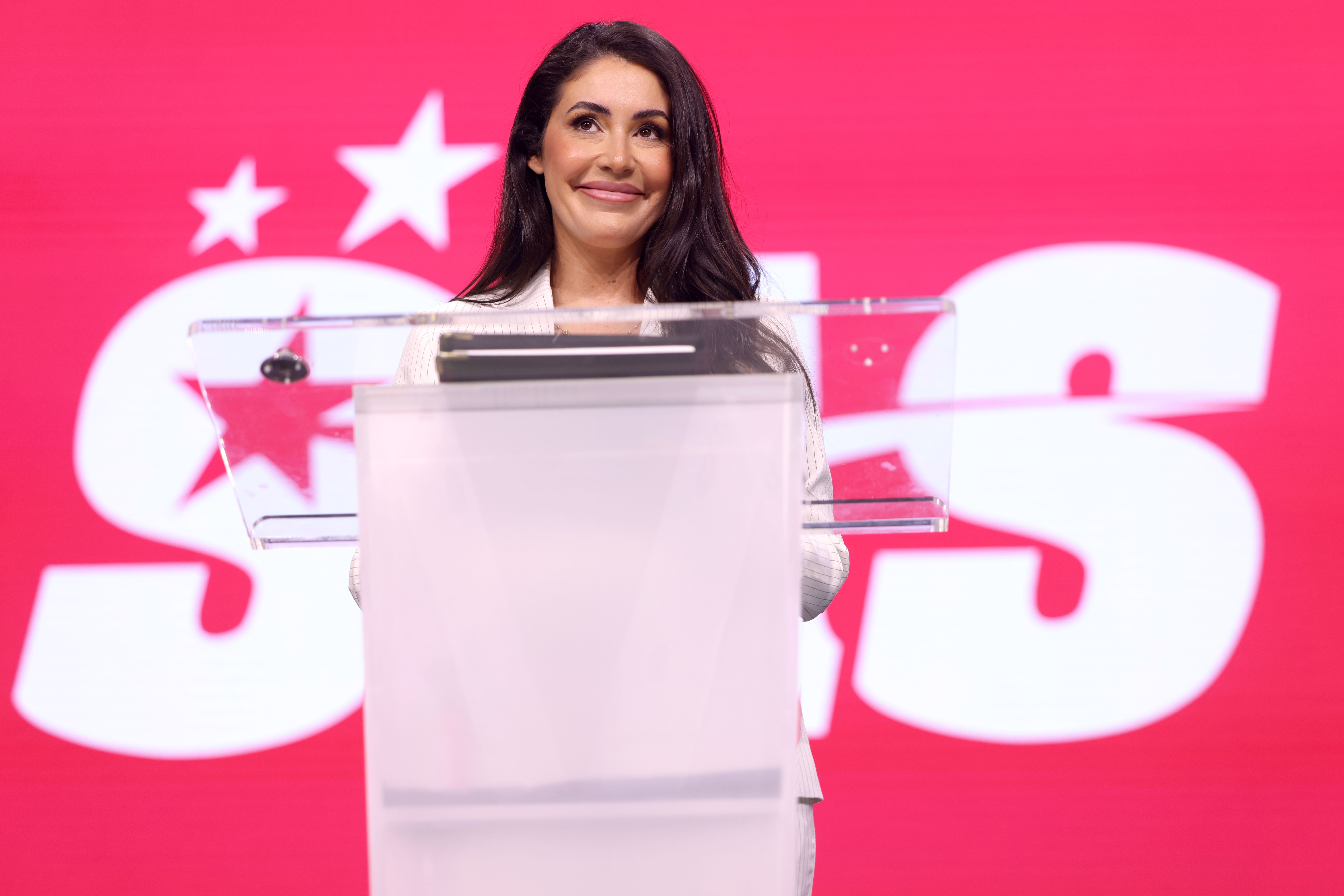 Woman in white blazer speaking at a clear acrylic podium against a bright pink backdrop.