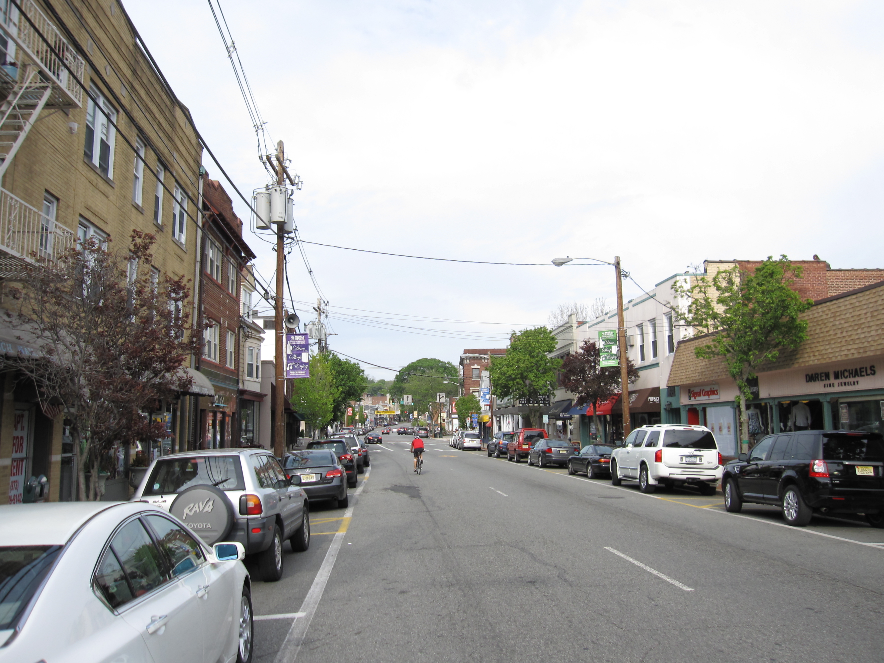 A commercial street with parked cars, storefronts, and utility poles on both sides.