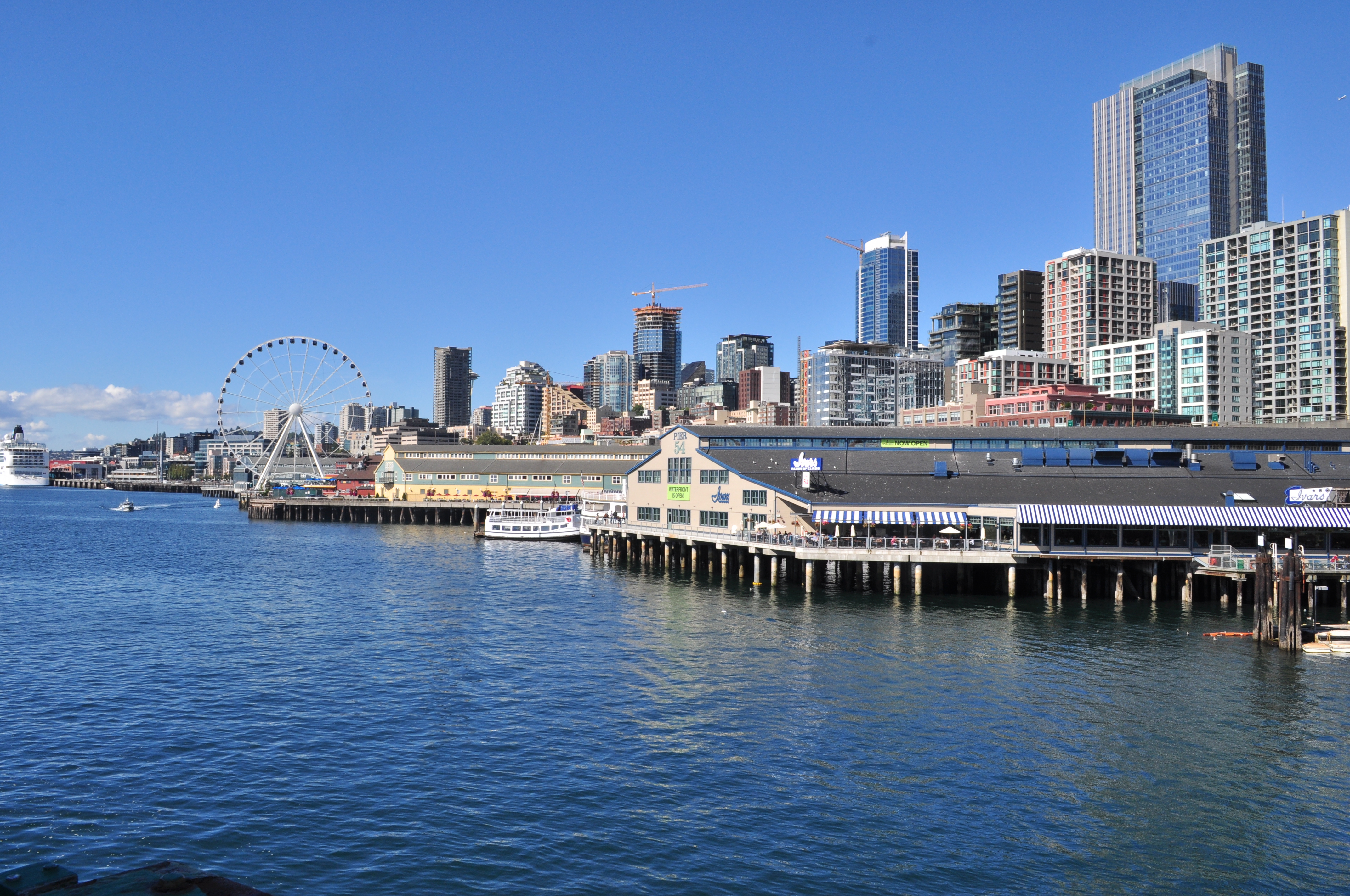 Seattle waterfront with historic piers, the Great Wheel, and downtown skyscrapers on a clear day.