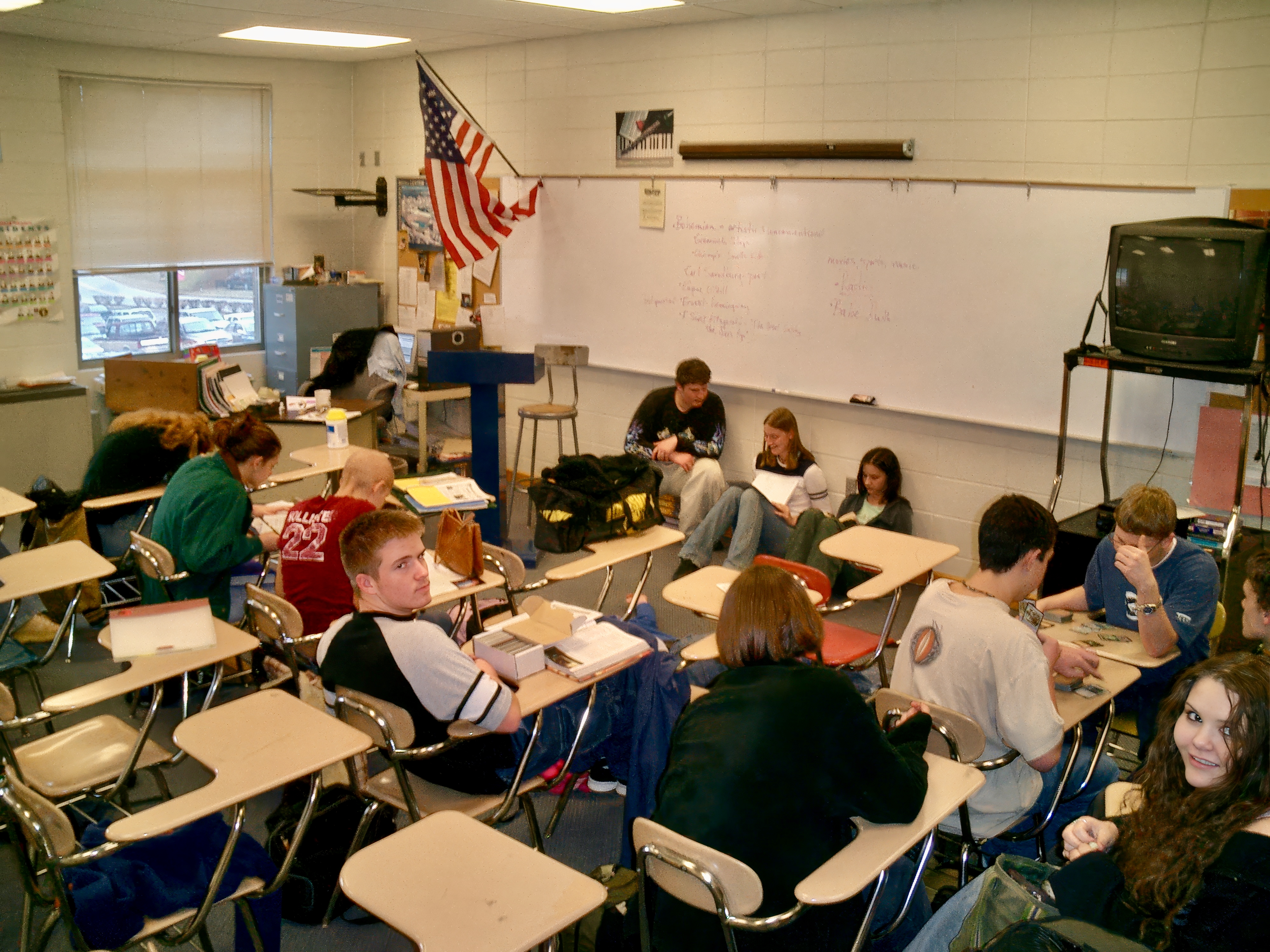 Students seated at desks in a full high school classroom with a whiteboard and American flag.